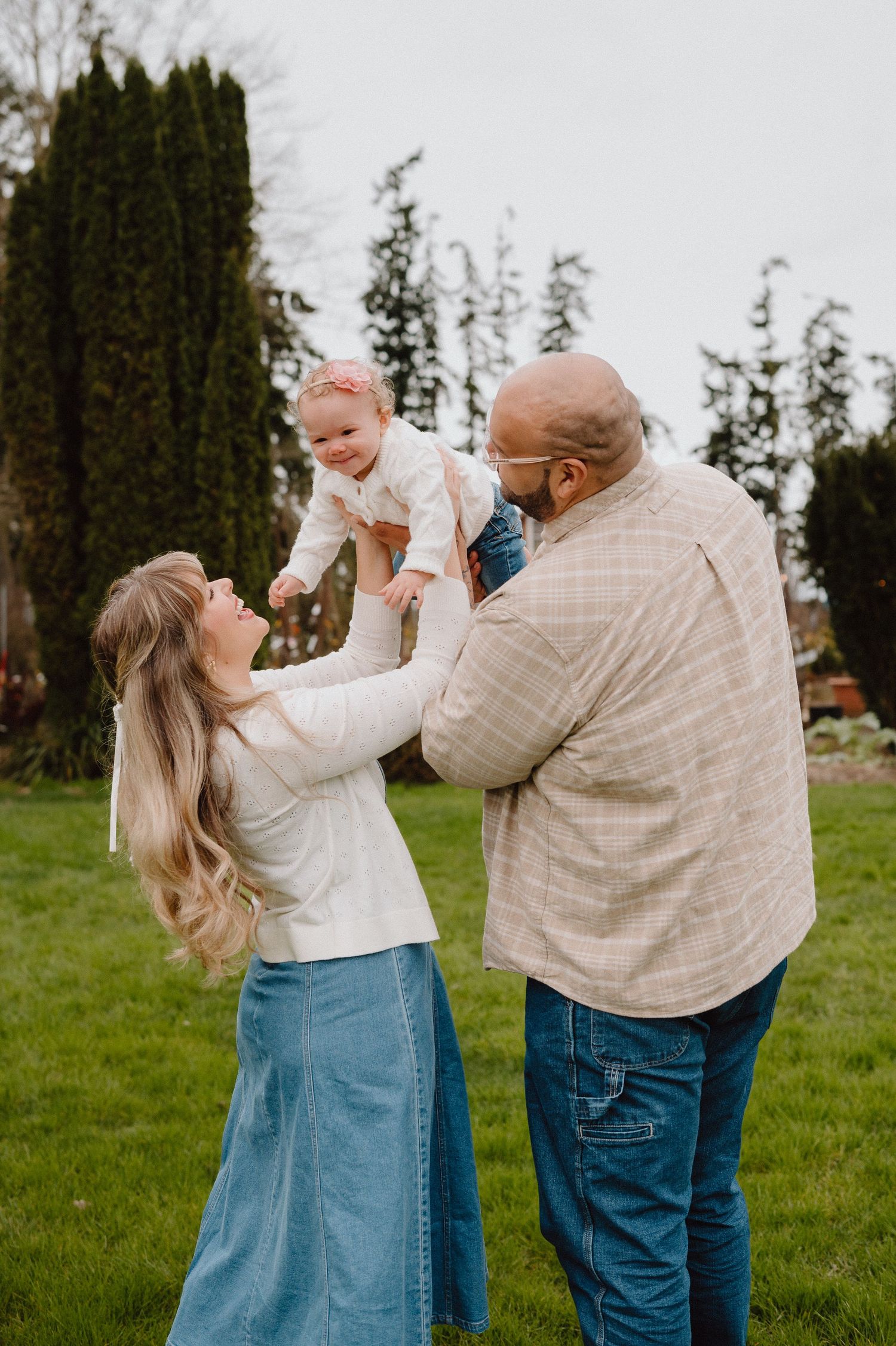 A couple joyfully lifts their smiling baby girl outdoors on a green lawn with tall evergreen trees in the background.