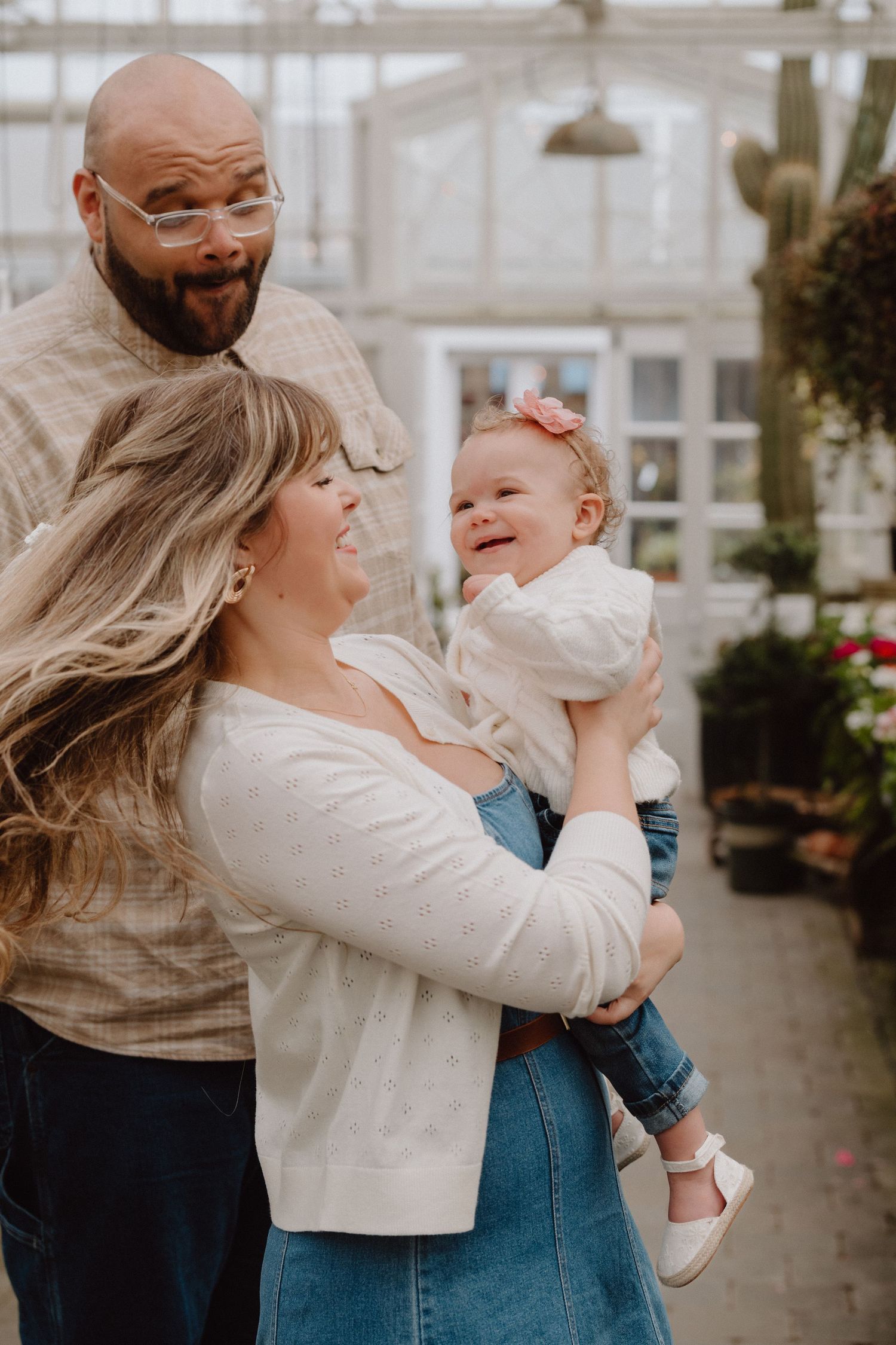 A joyful family with a bald father, blonde mother, and laughing baby girl in a greenhouse setting.