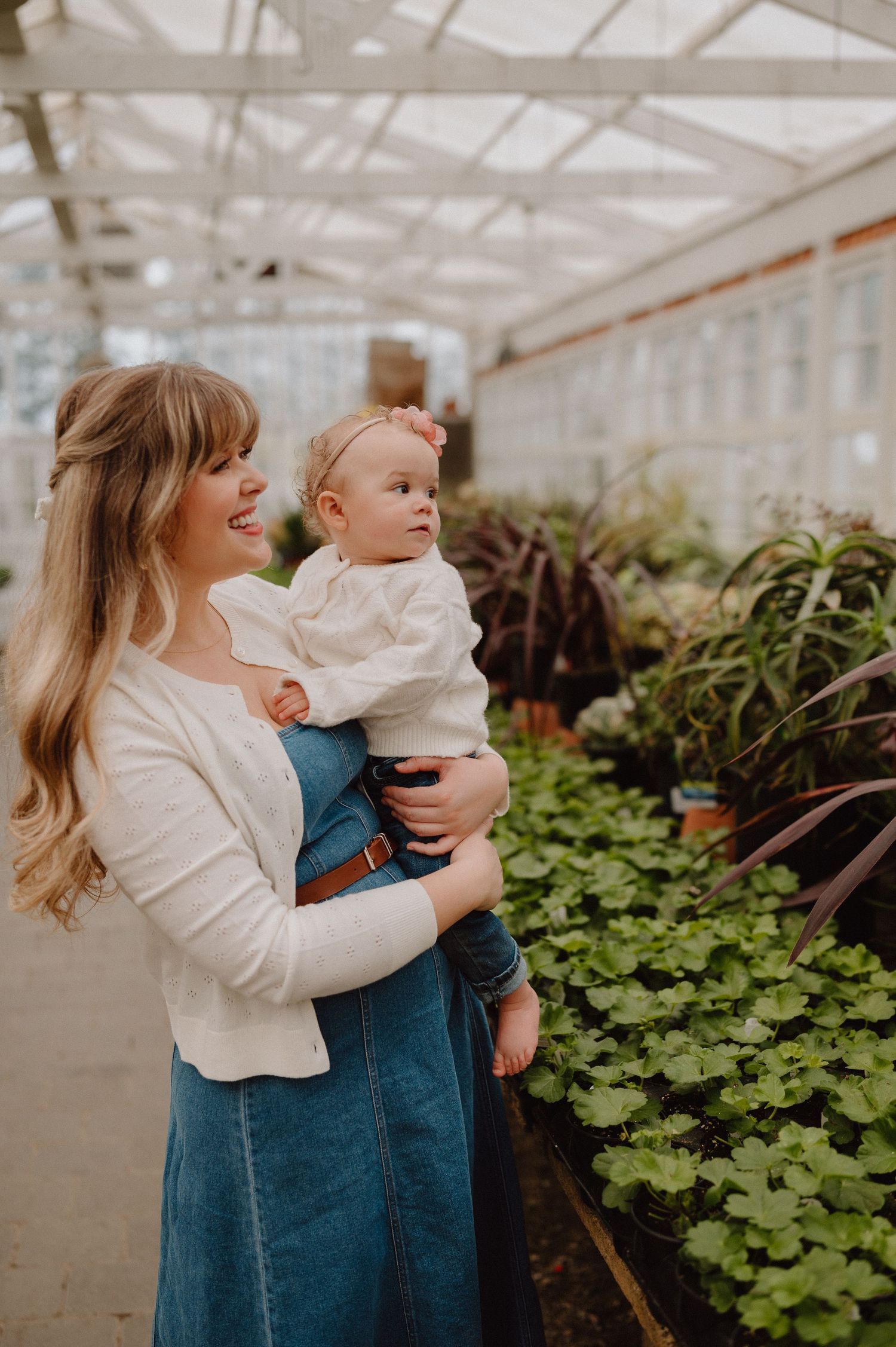 A smiling mother holds her baby girl while browsing green plants in a bright greenhouse.