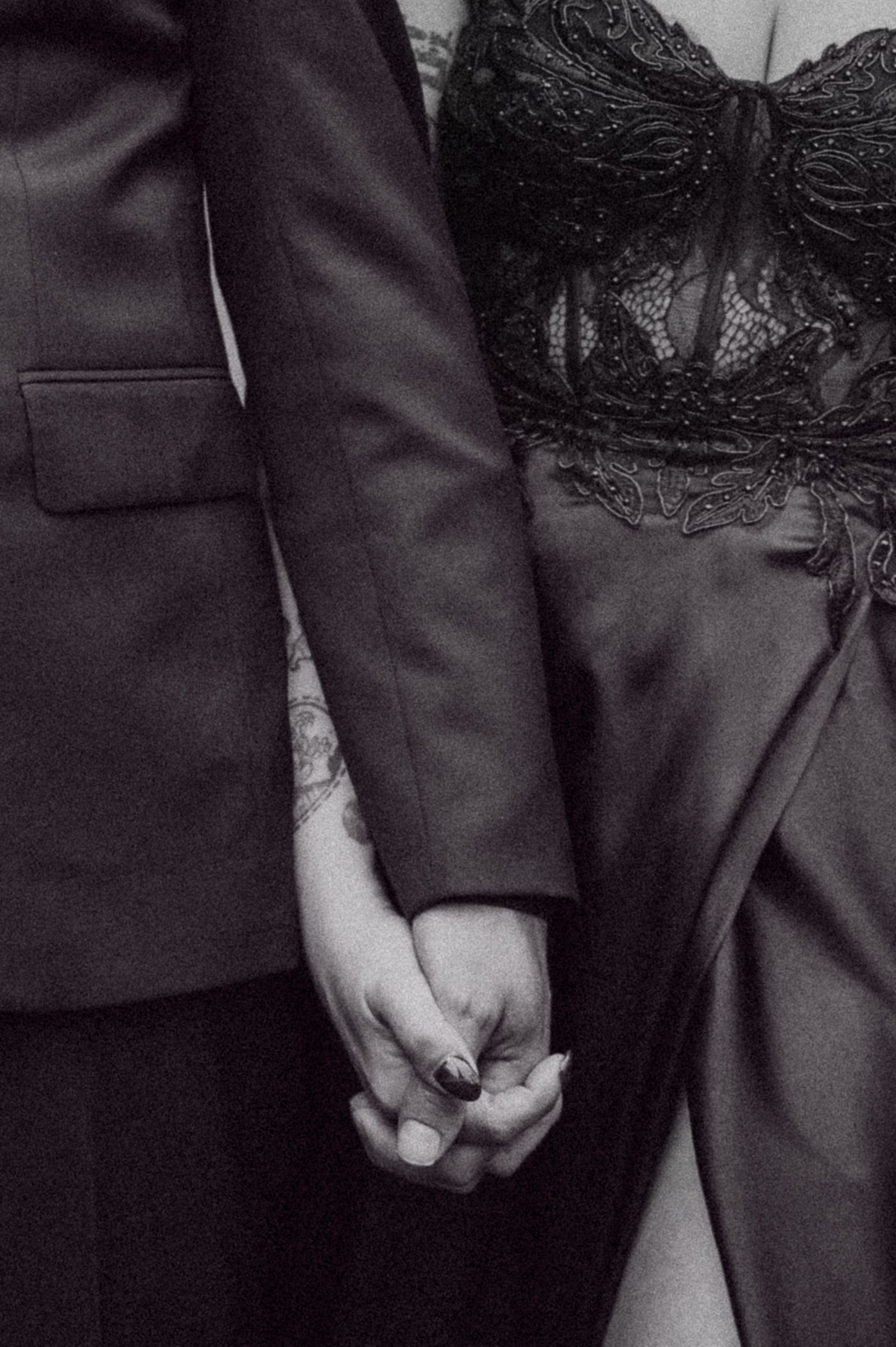 Black and white photo of a couple holding hands, man in suit with tattoos, woman in black lace dress.