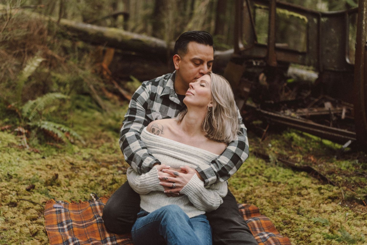 Couple embracing on plaid blanket in mossy forest, man kissing woman's head, wearing cozy sweaters.