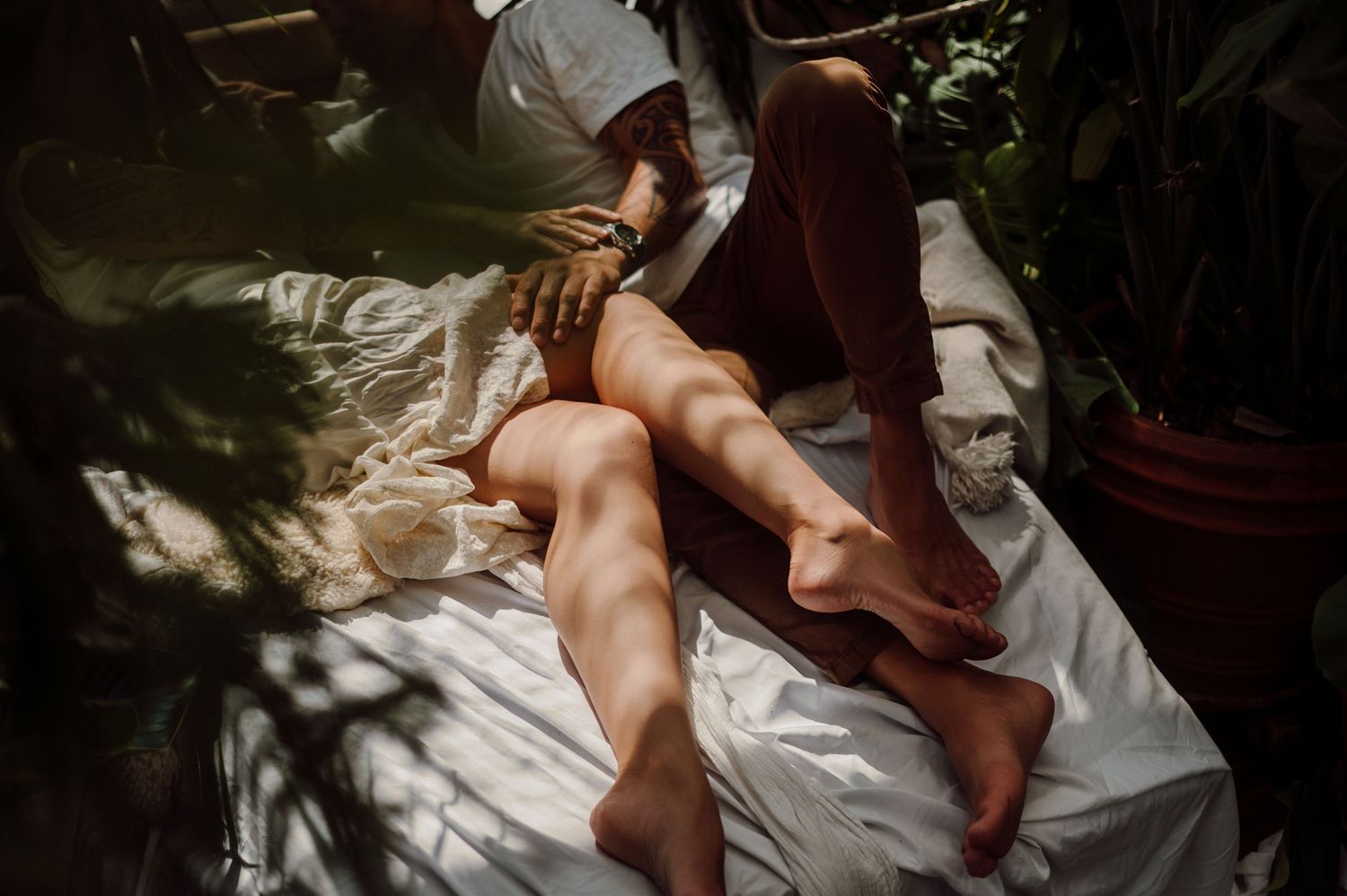 A couple embraces intimately in bed with white sheets while natural light streams through the window.