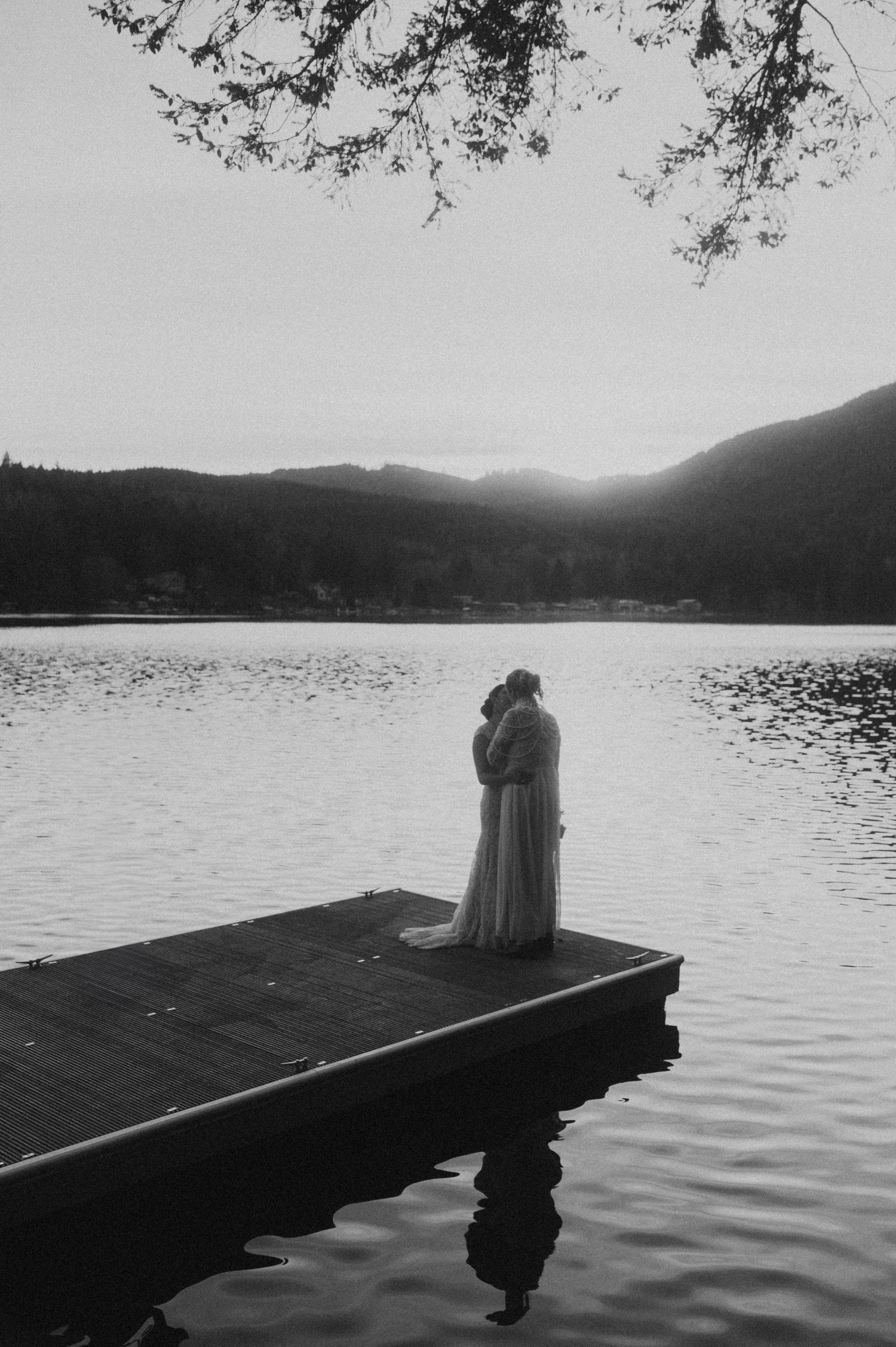 A silhouetted figure sits contemplatively on a wooden dock overlooking a serene lake with mountains in the background.