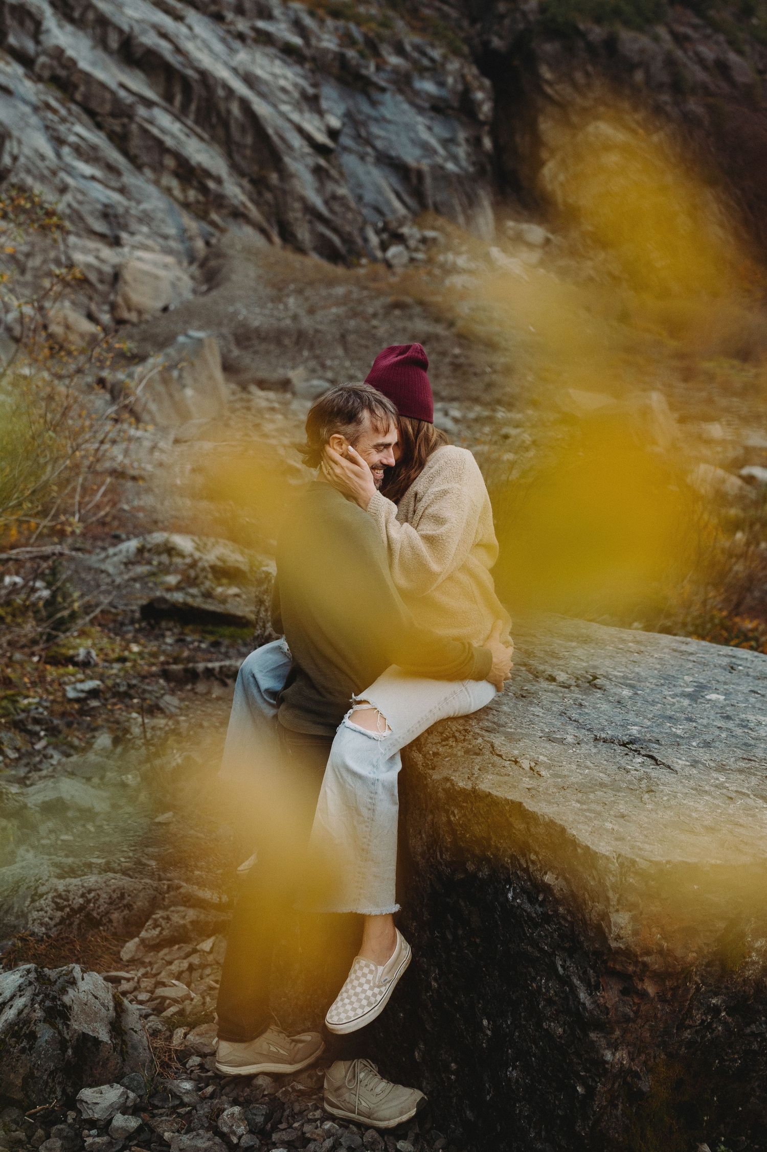 A couple embraces against a rocky cliff wall with yellow autumn foliage in the foreground creating a dreamy atmosphere.