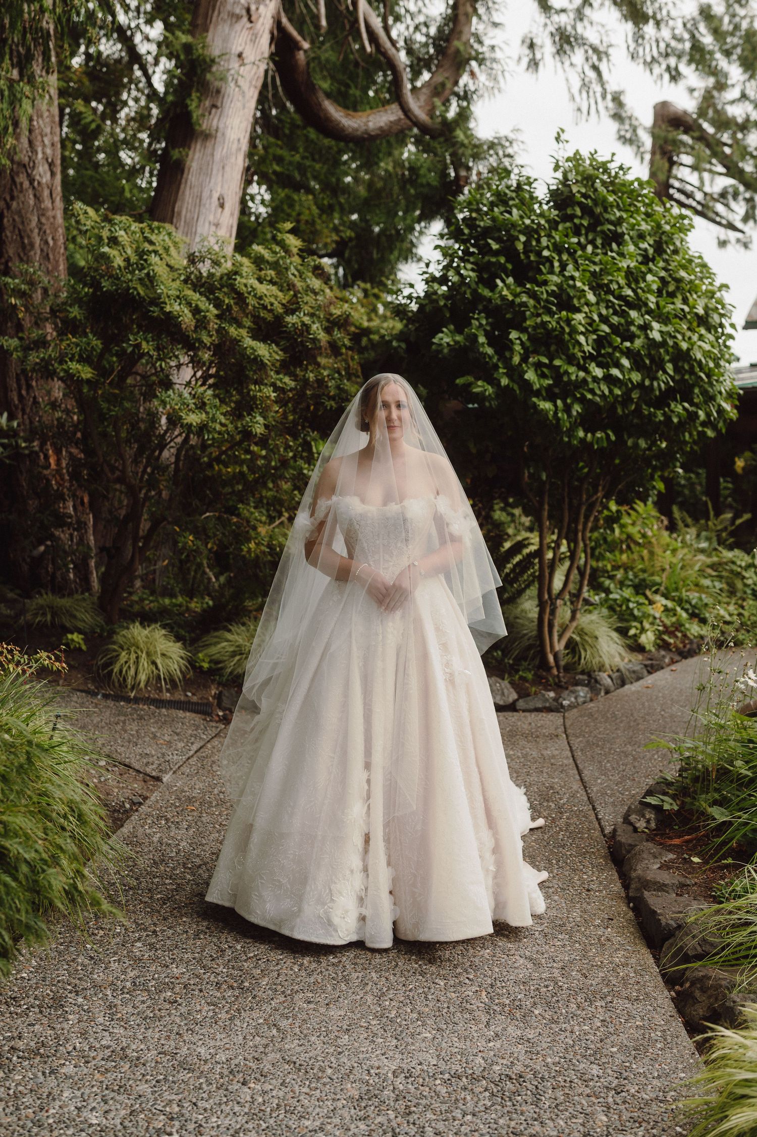 A bride in a flowing white ballgown wedding dress and veil stands on a gravel path surrounded by lush garden foliage.