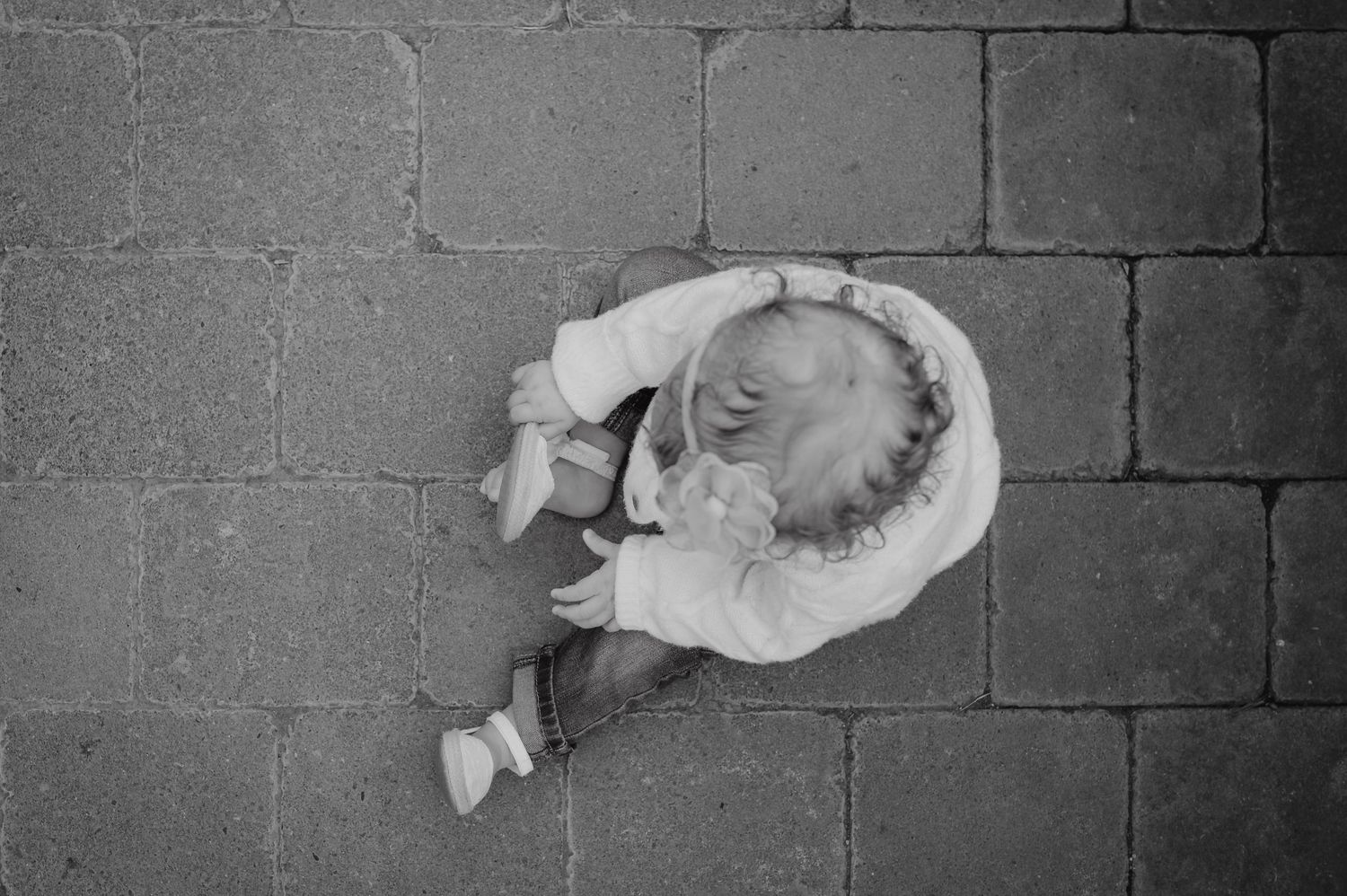 Black and white aerial view of a small child sitting on cobblestone pavement, wearing a white dress and shoes.