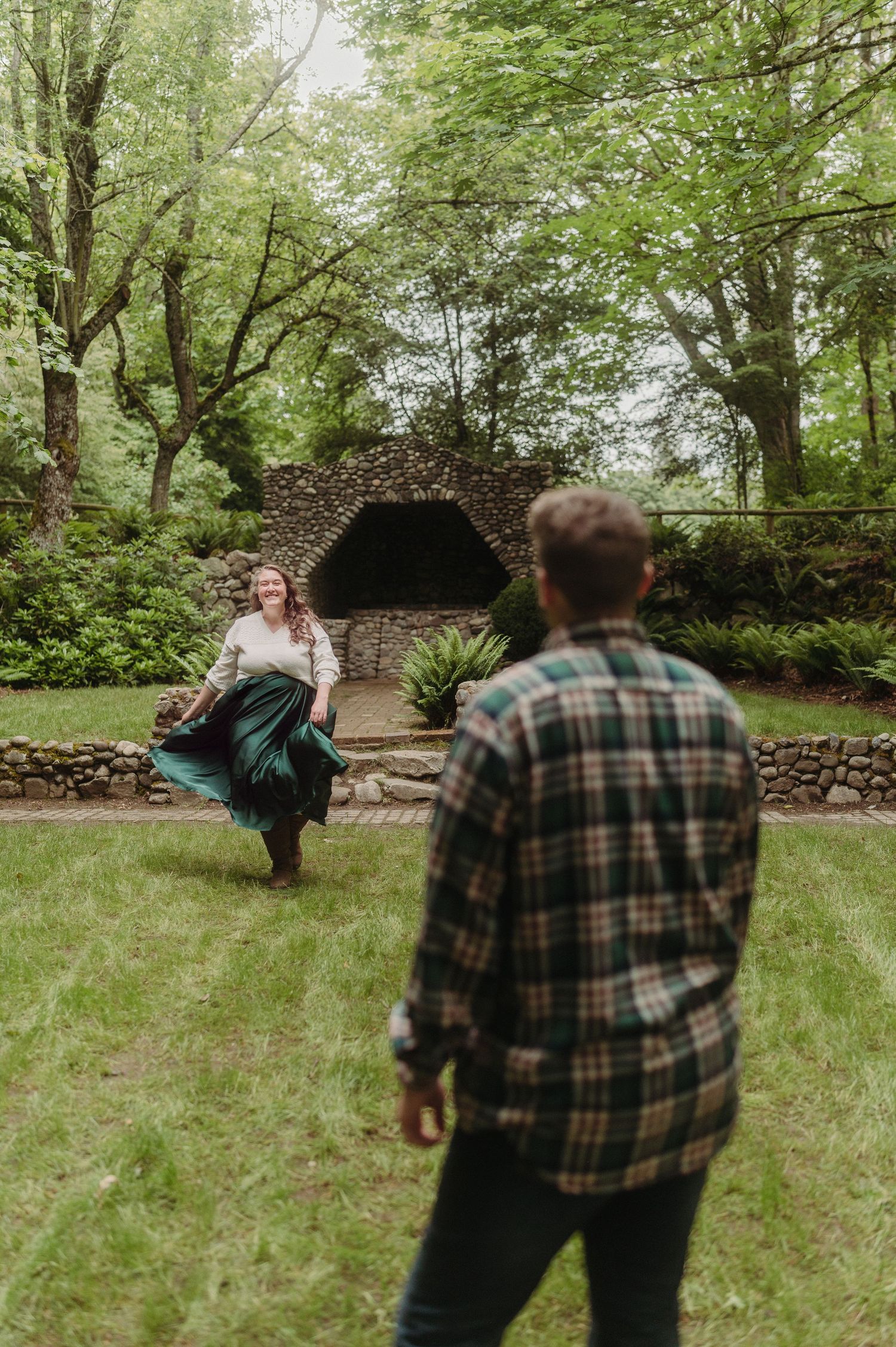 A couple plays in a lush garden, woman twirling green skirt near stone grotto while man in plaid shirt watches.