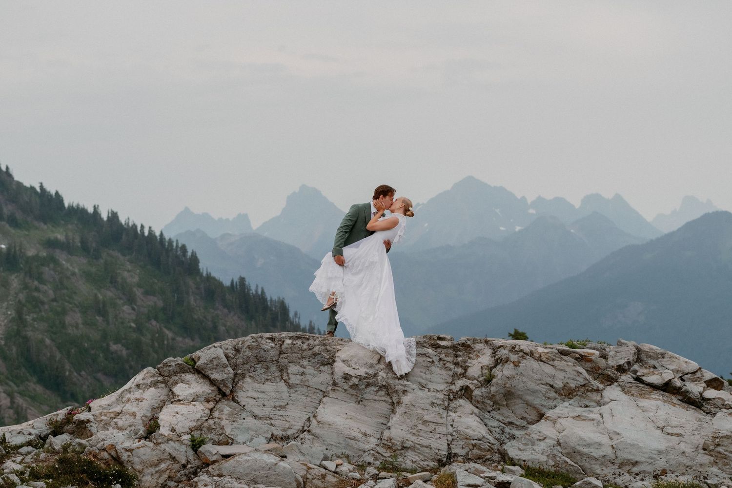 A couple in wedding attire stands atop a dramatic mountain cliff overlooking majestic peaks in the background.