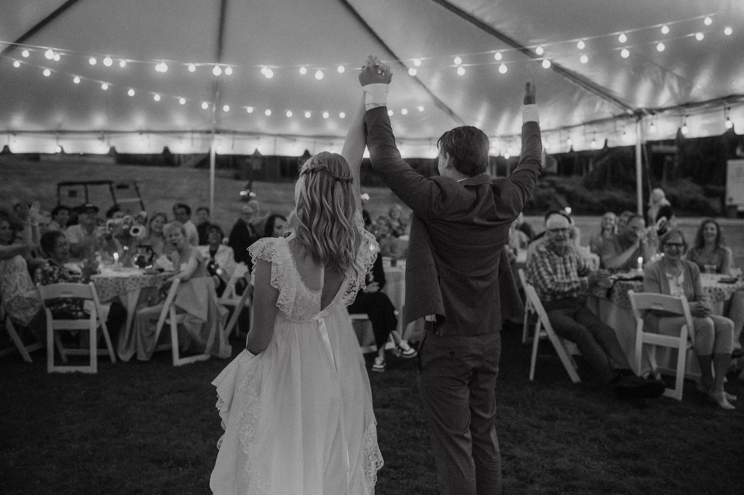 A couple dances under string lights at an outdoor wedding reception with white folding chairs visible in the background.