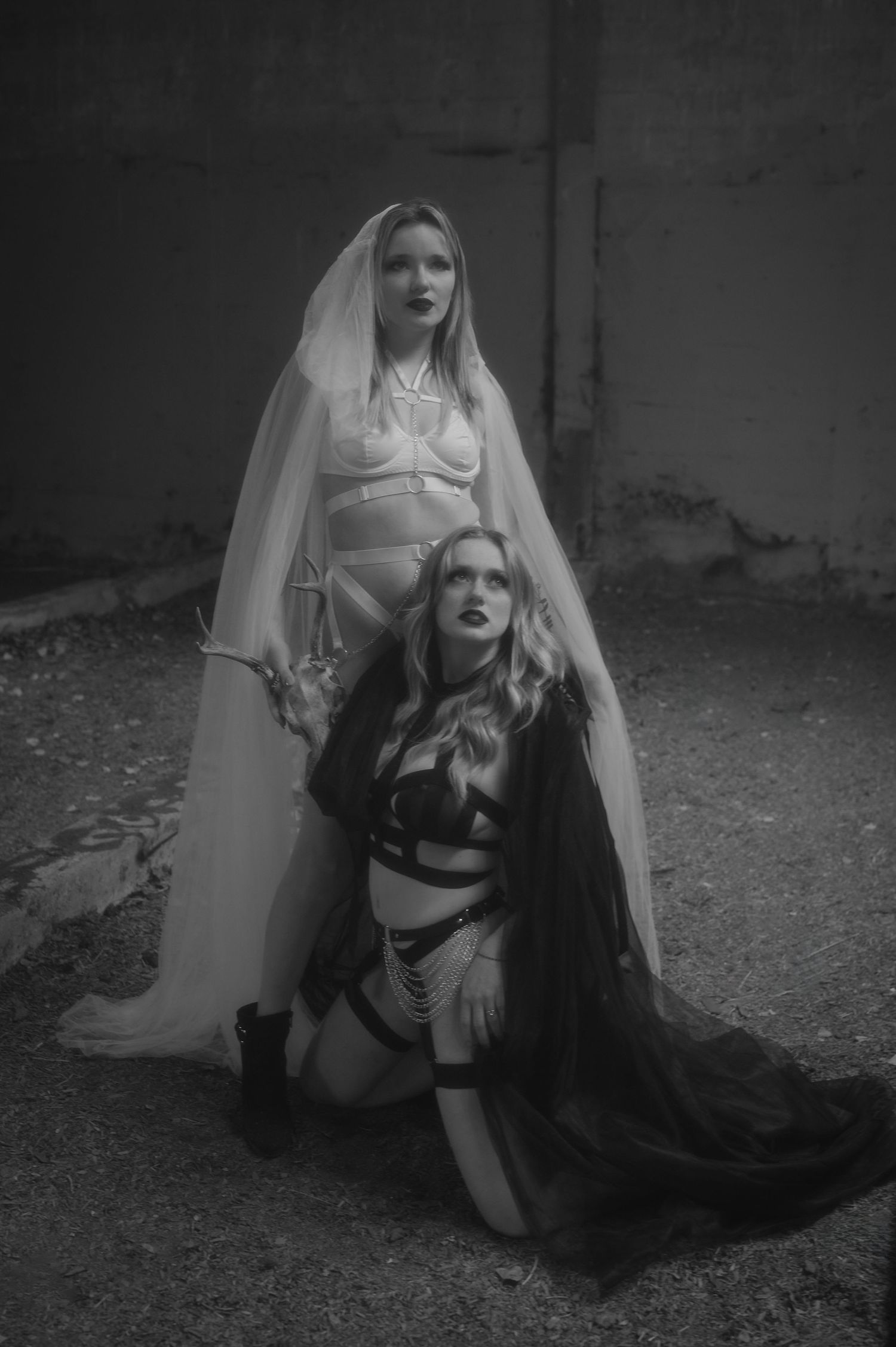 Two figures in gothic-inspired outfits pose dramatically in black and white, wearing veils and dark clothing in a moody setting.