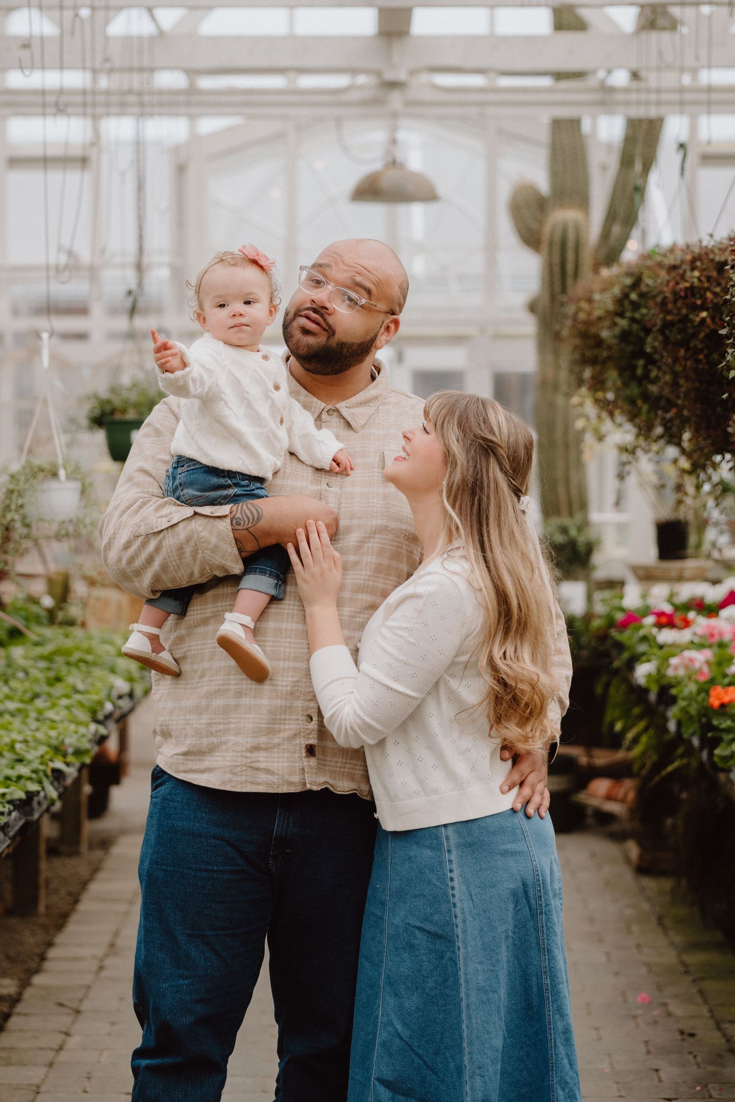 A family poses in a lush greenhouse, father holding baby girl while mother gazes up lovingly in neutral tones.