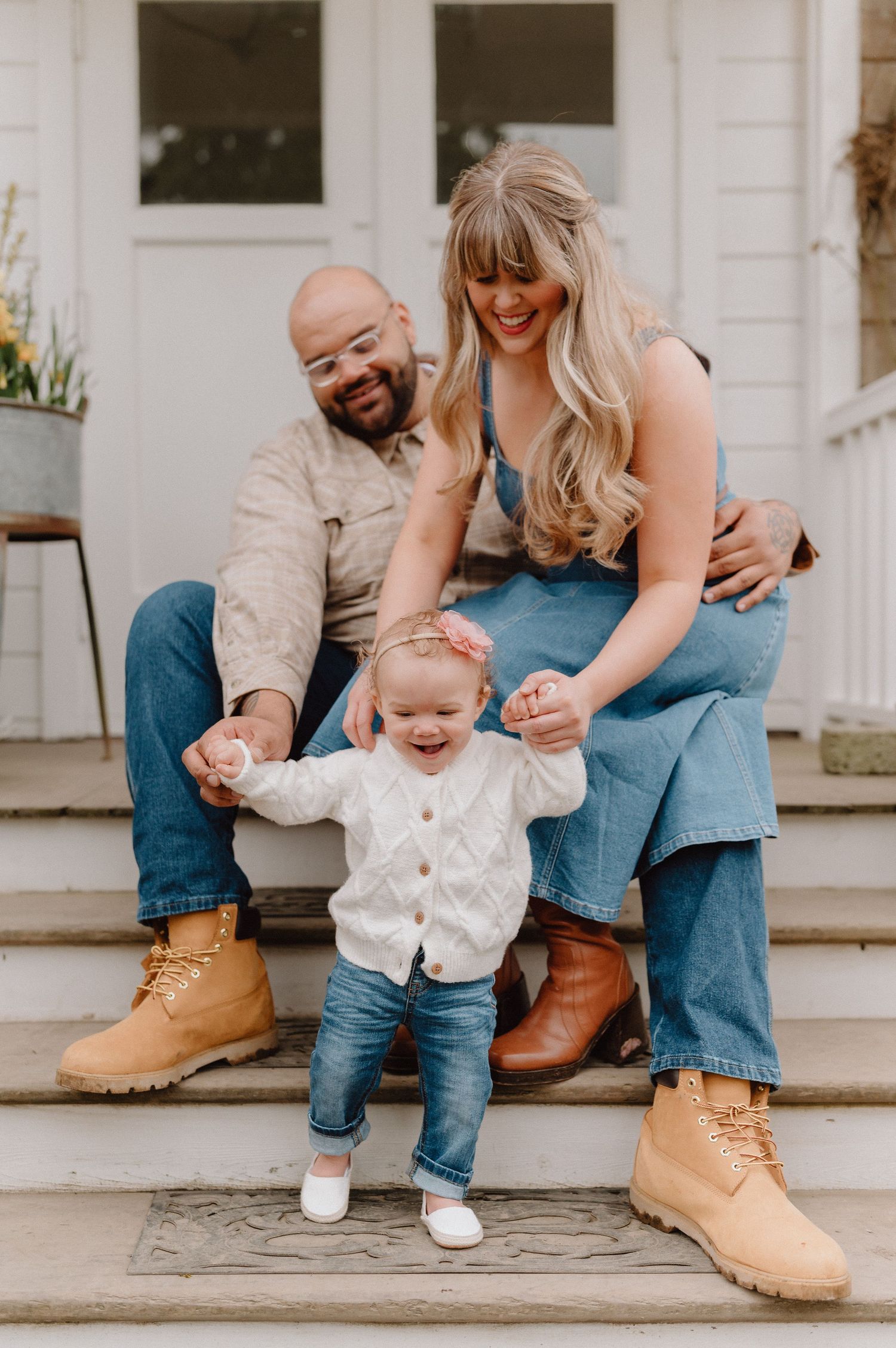 Happy family with bald dad, blonde mom in denim, and toddler girl sitting on porch steps holding hands.