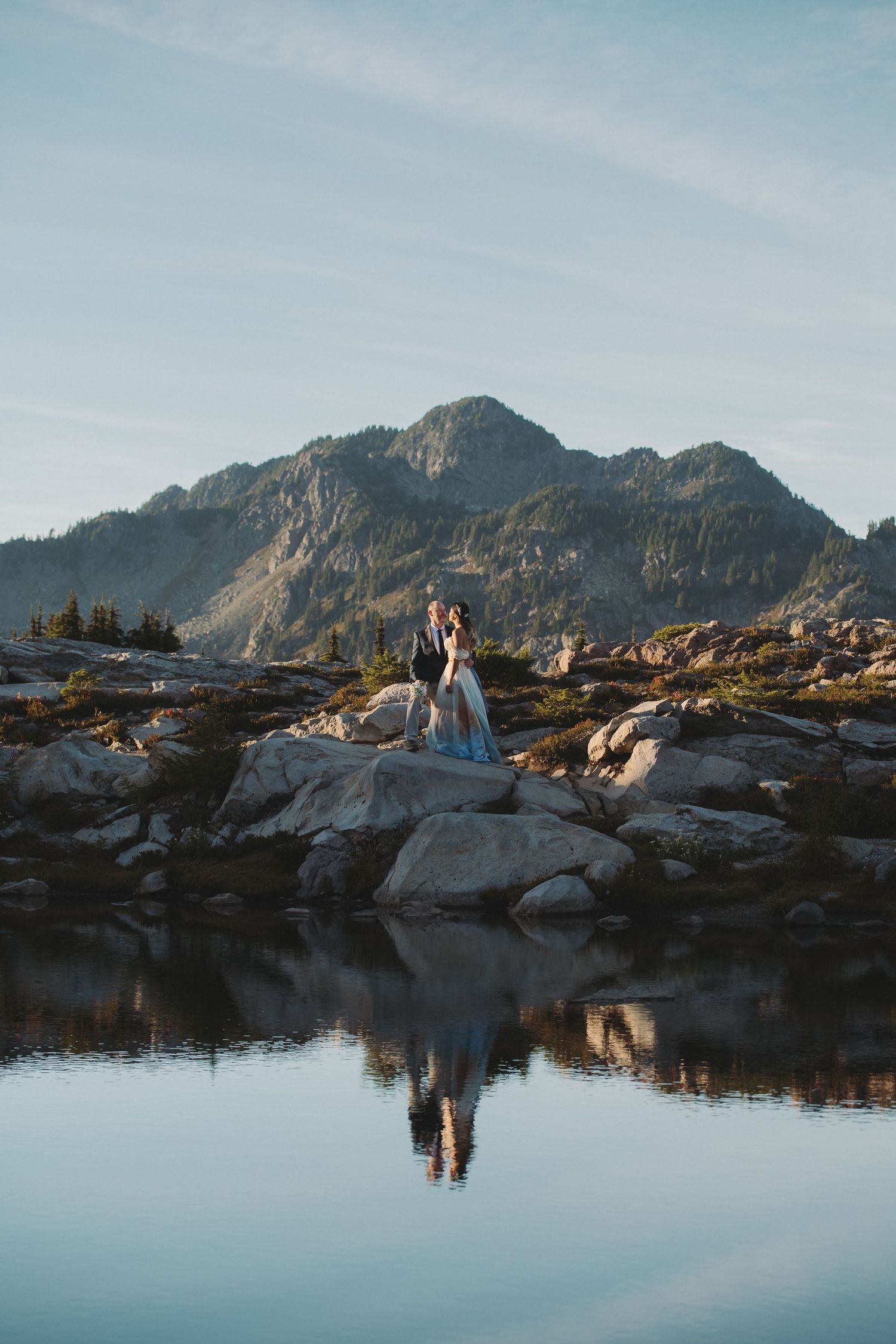 Two people embrace on rocky terrain near a calm mountain lake with perfect reflection during golden hour.
