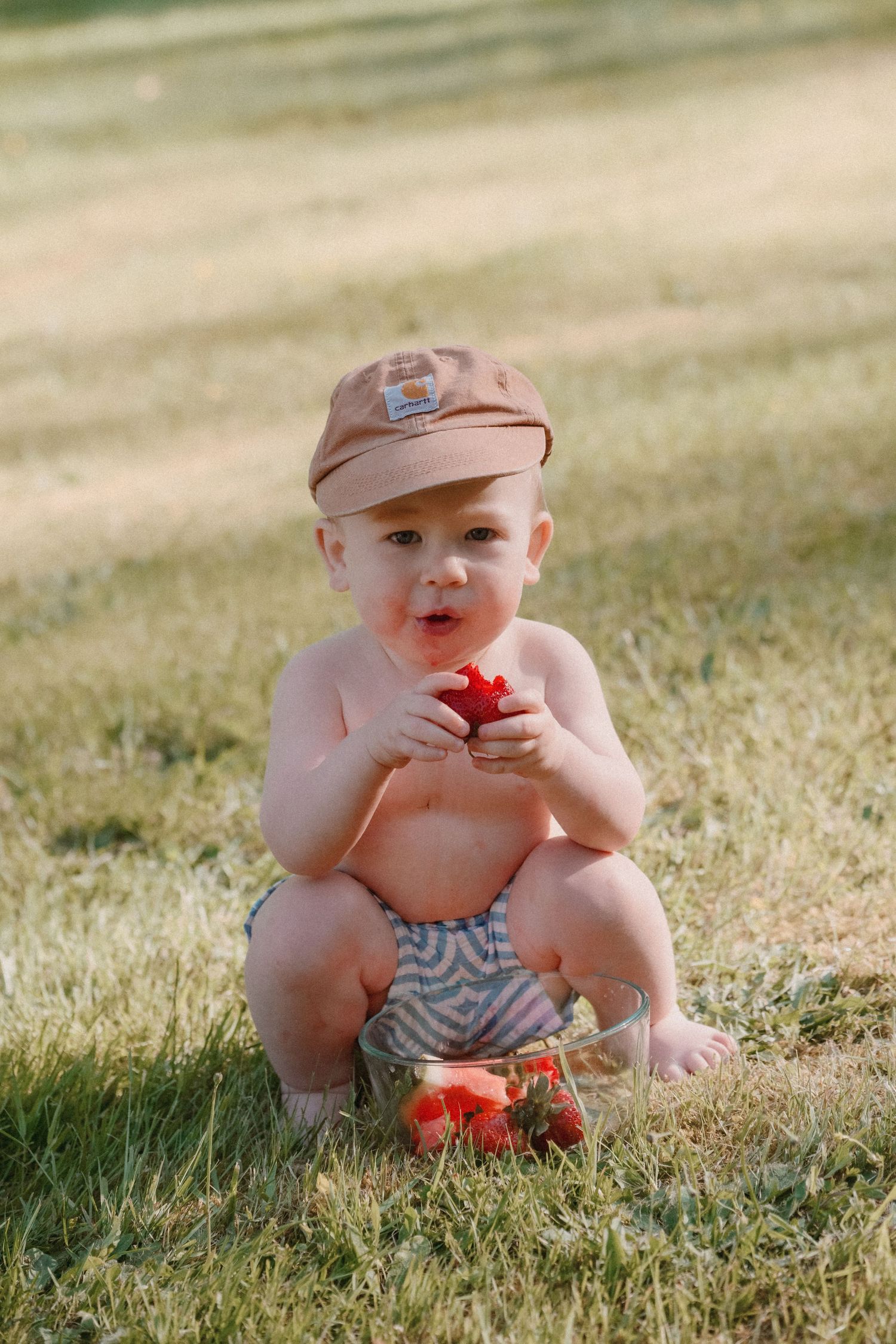Baby wearing newsboy cap sitting in grass playing with red tomatoes on sunny summer day.