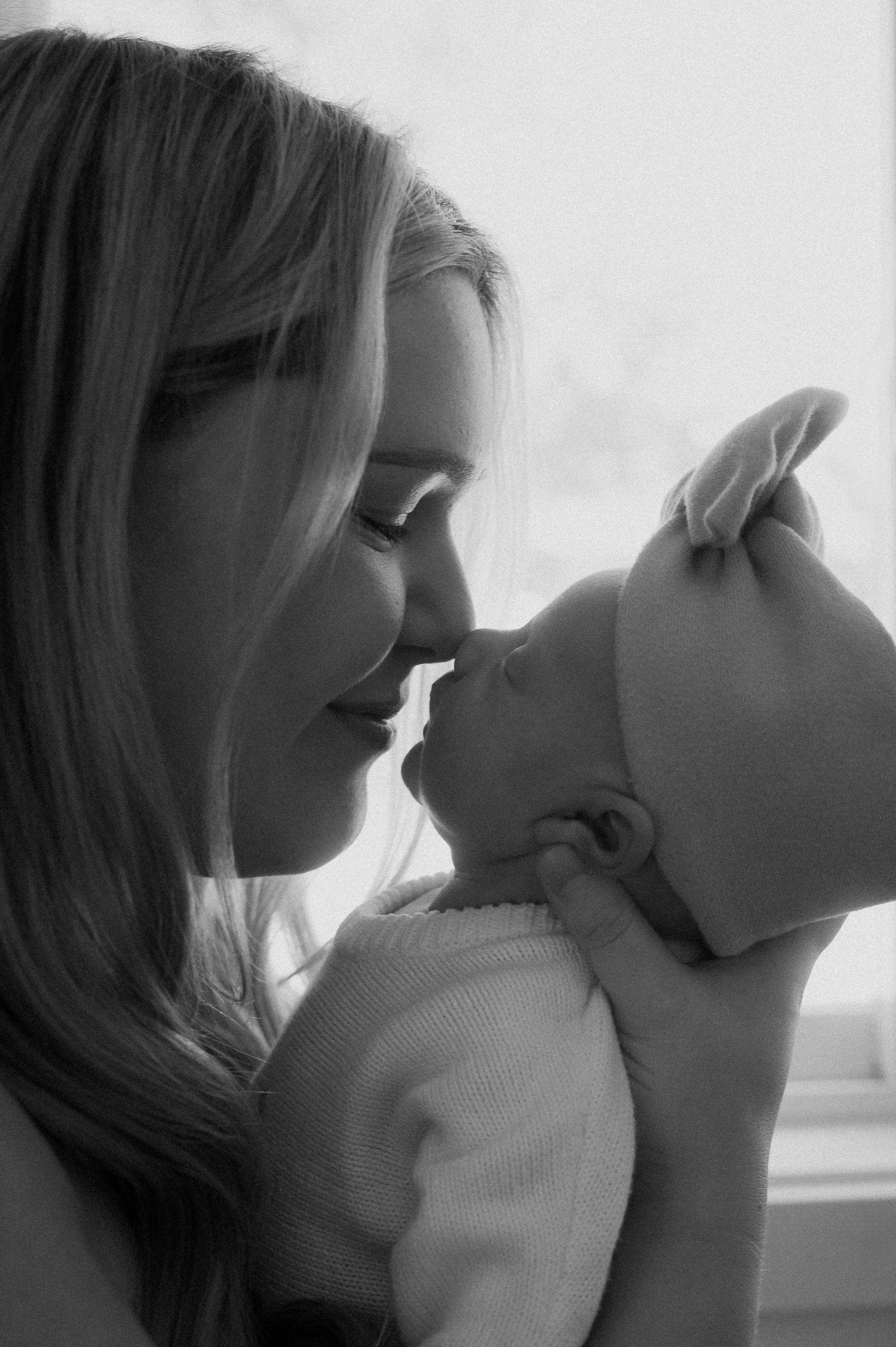 A tender black and white portrait of a woman embracing and nuzzling a small baby in soft lighting.