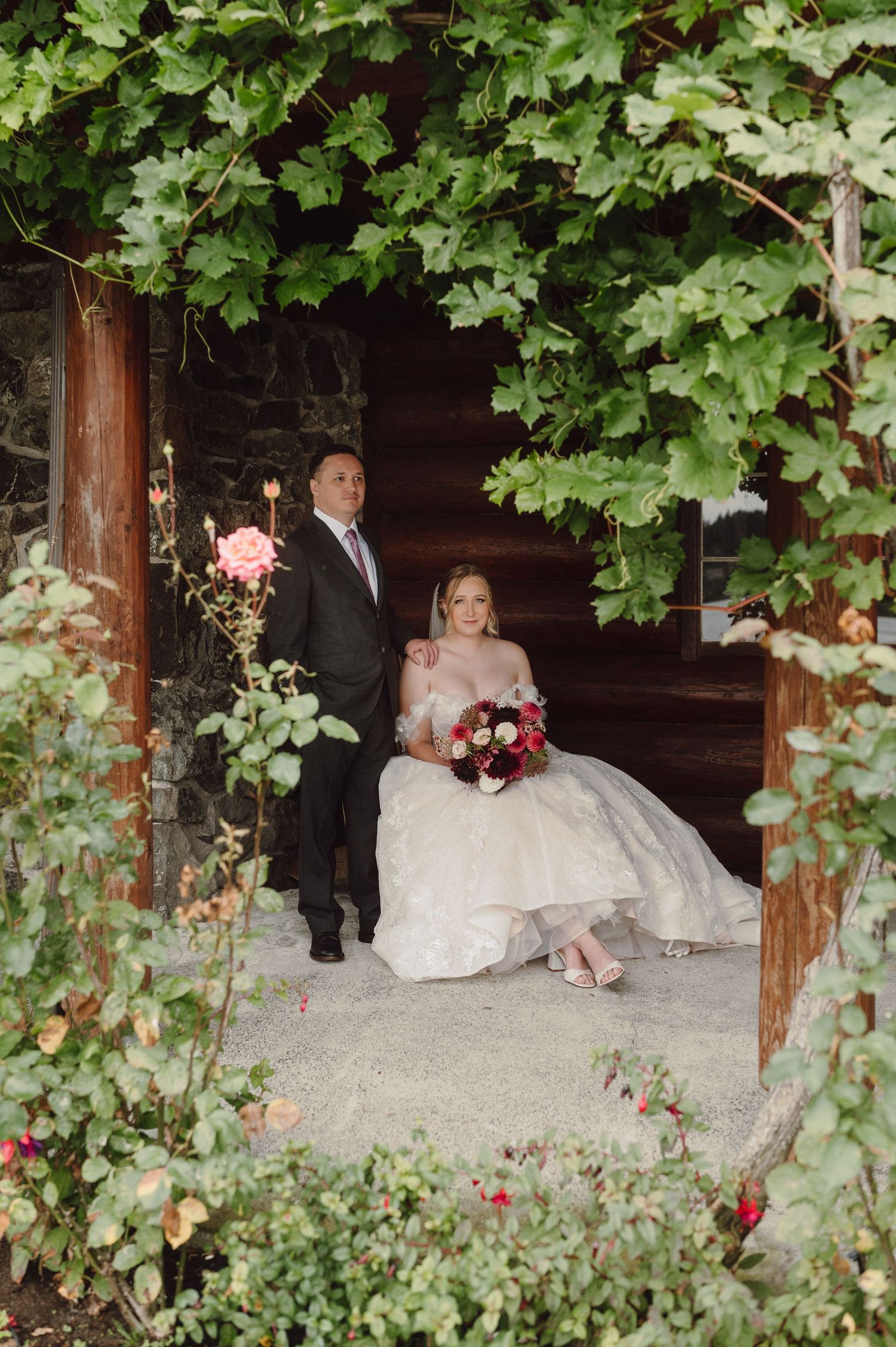 A couple sits on a rustic wooden bench surrounded by lush green ivy and garden foliage at an outdoor venue.