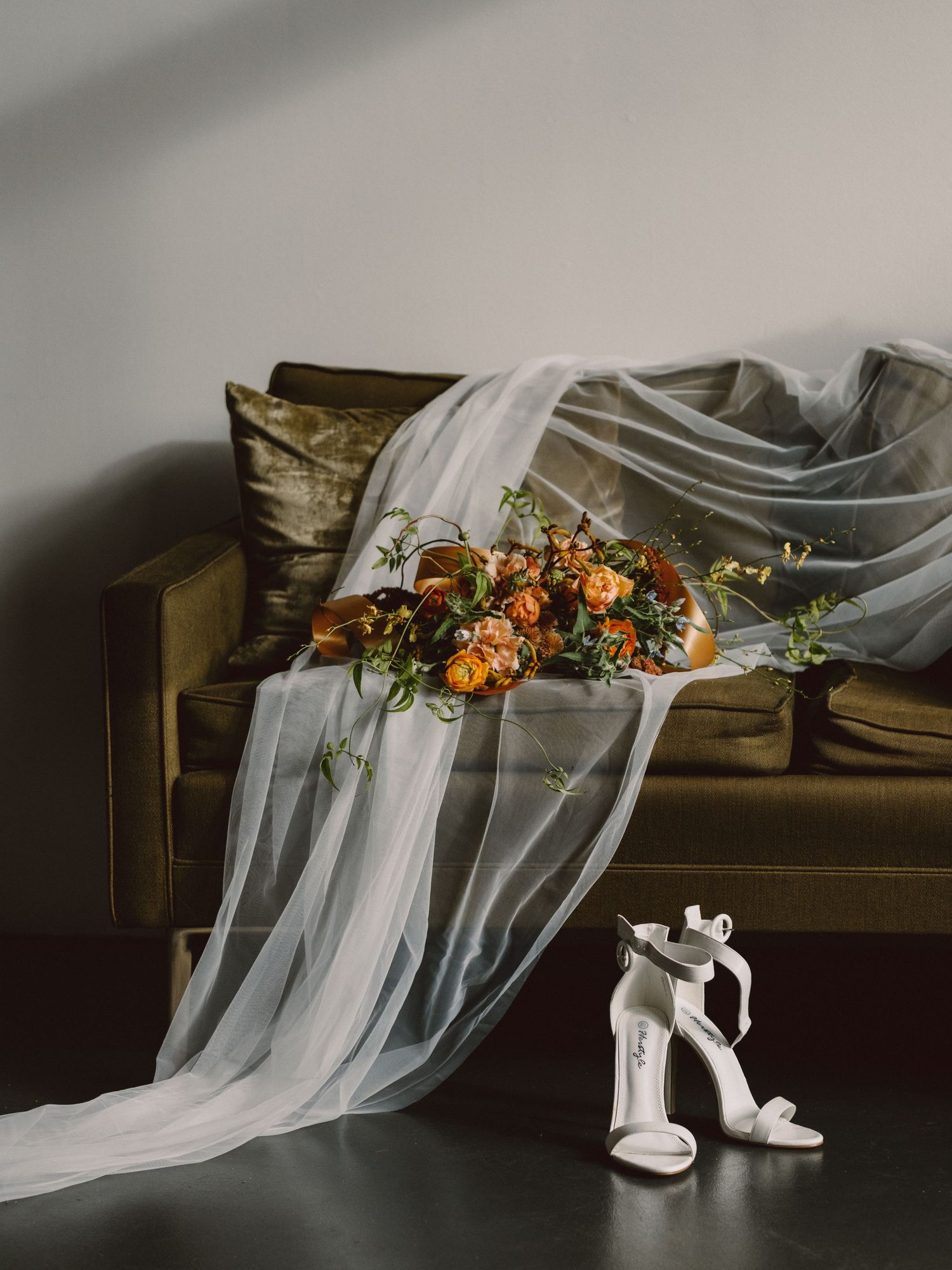 A delicate white wedding veil drapes elegantly across a vintage couch with orange floral accents and white bridal shoes.