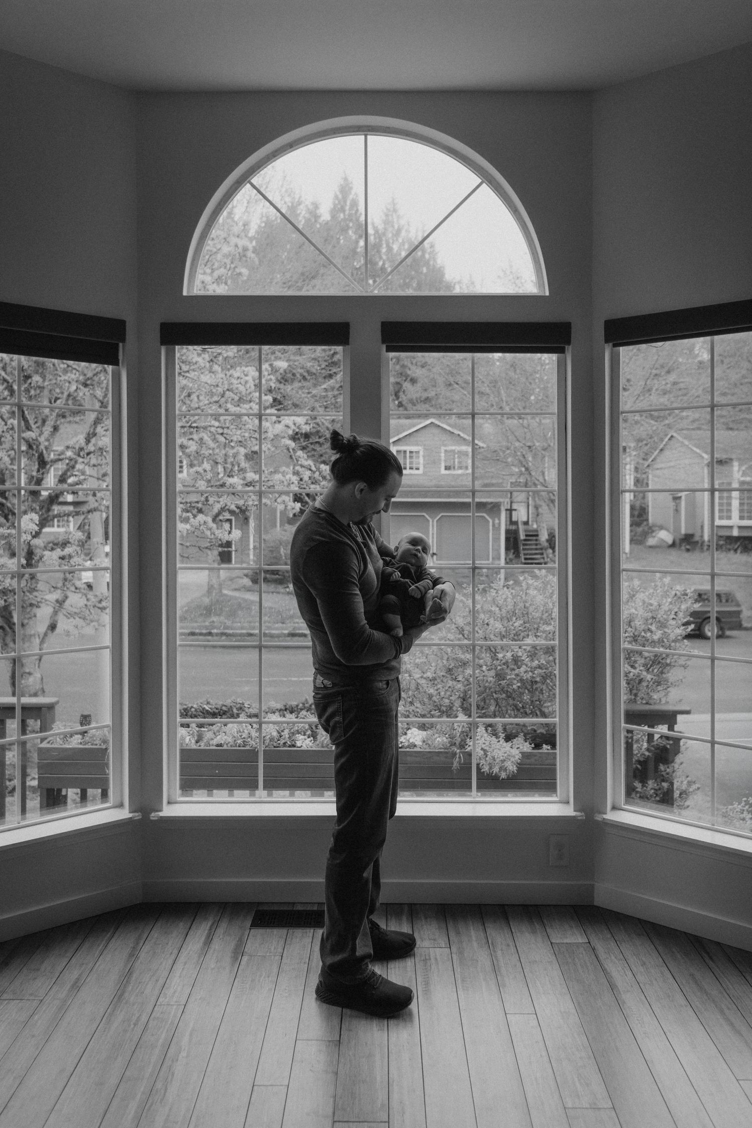 Black and white photo of a person holding a baby while standing in a bay window overlooking a suburban neighborhood.
