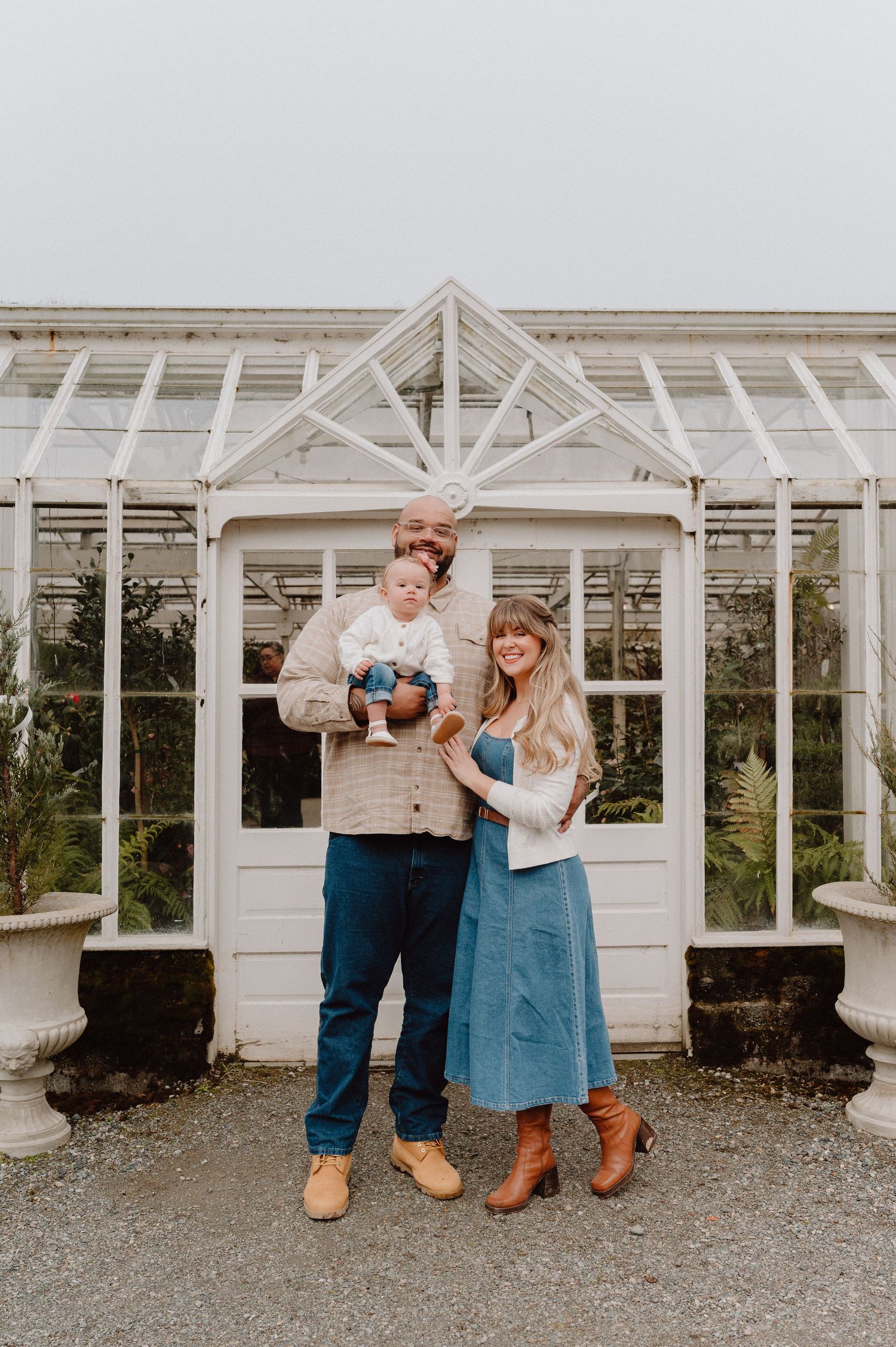 A smiling couple stands before a white greenhouse, holding their baby, dressed in casual fall outfits.