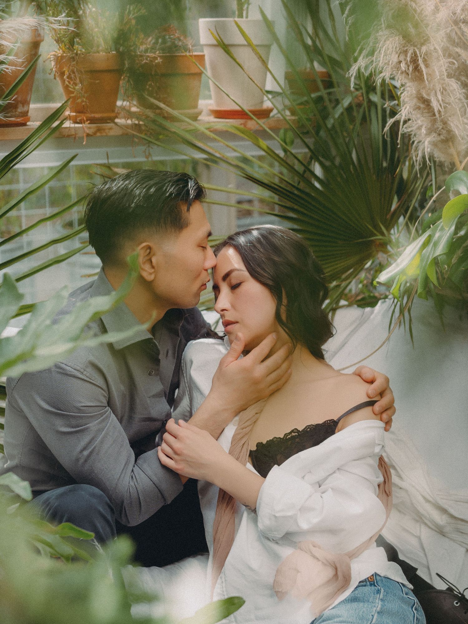 A romantic embrace between a couple surrounded by lush greenery and potted plants in a greenhouse setting.