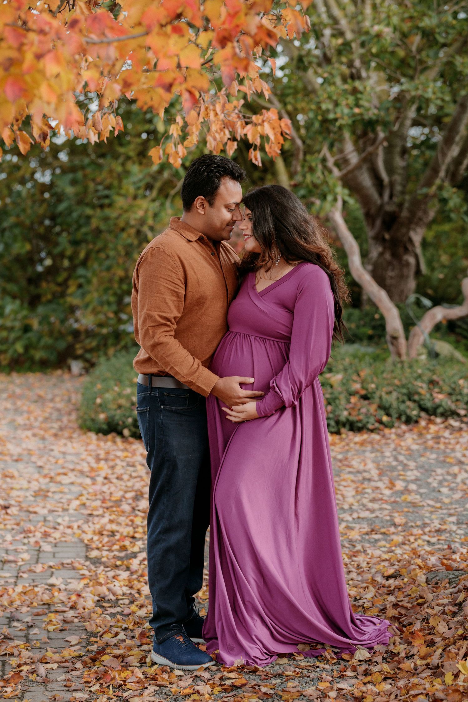 A couple embraces under vibrant orange fall foliage wearing burgundy and brown clothing.