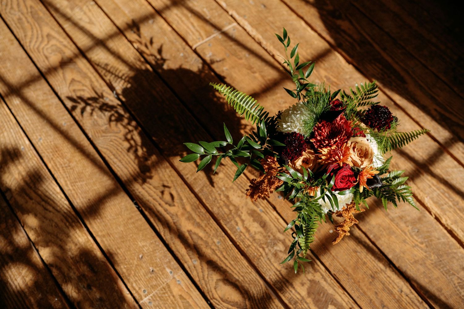 Wedding bouquet with red roses and greenery laying on wooden floor with dramatic diagonal shadows.