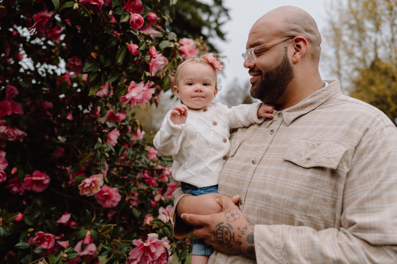Bald father with glasses and bow tie holds baby near pink flowering bush outdoors.
