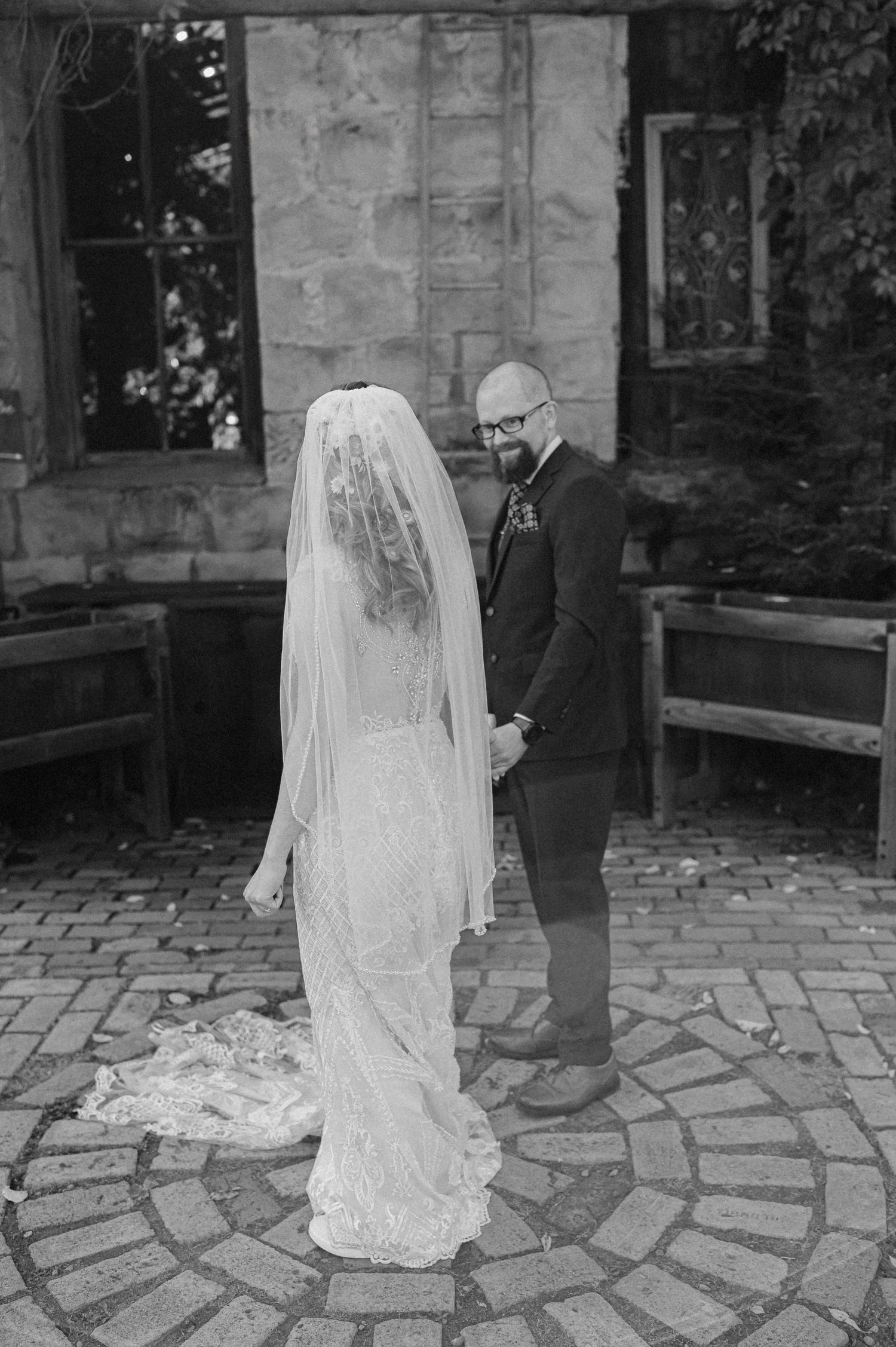 A black and white wedding photo showing a couple on a stone patio with the bride wearing a long flowing veil and lace gown.