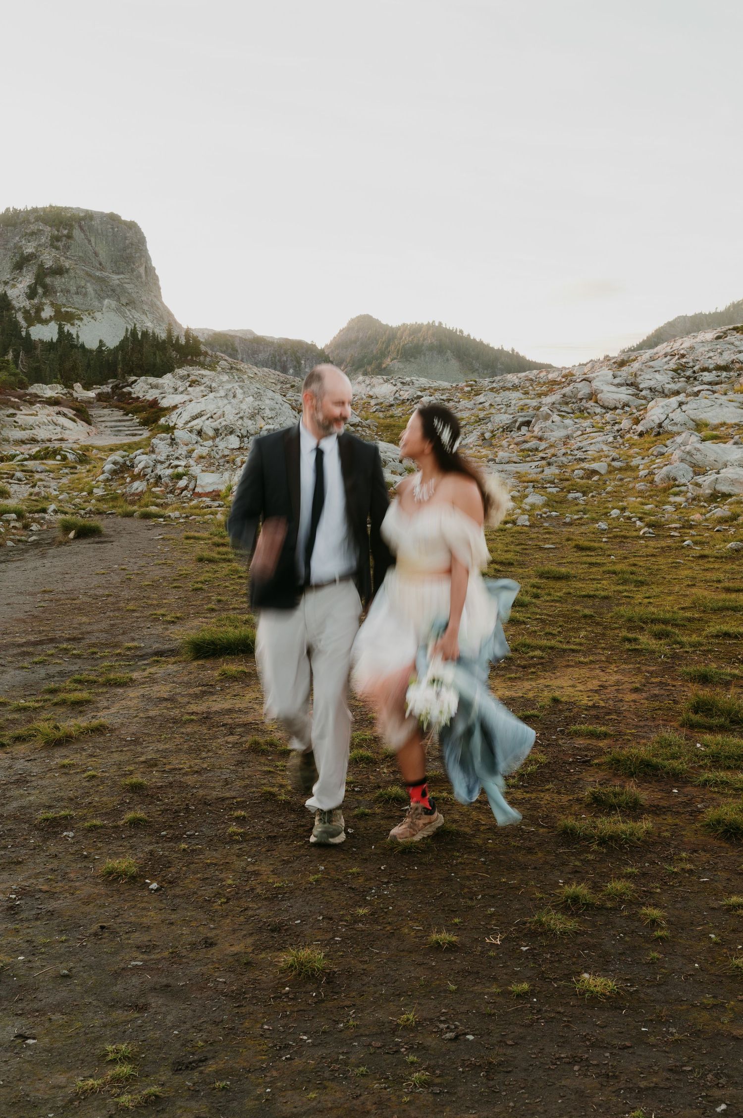 Couple in wedding attire walks hand-in-hand through a rocky mountain meadow at dusk, laughing together.