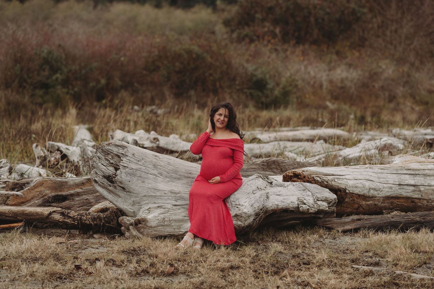 Pregnant woman in red off-shoulder gown poses on driftwood logs in a natural outdoor setting.