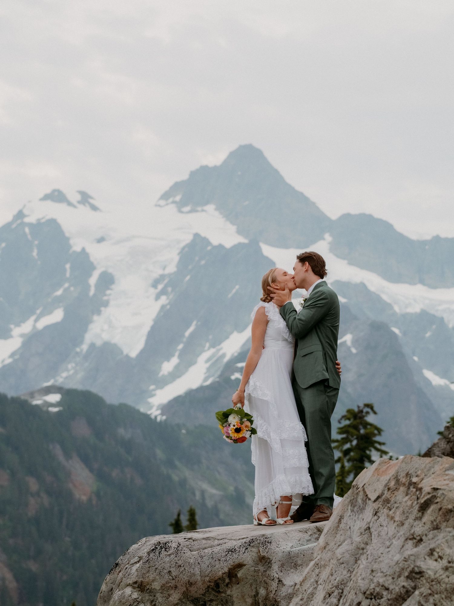 A couple shares a romantic moment on a rocky mountain peak with snow-capped mountains in the background.