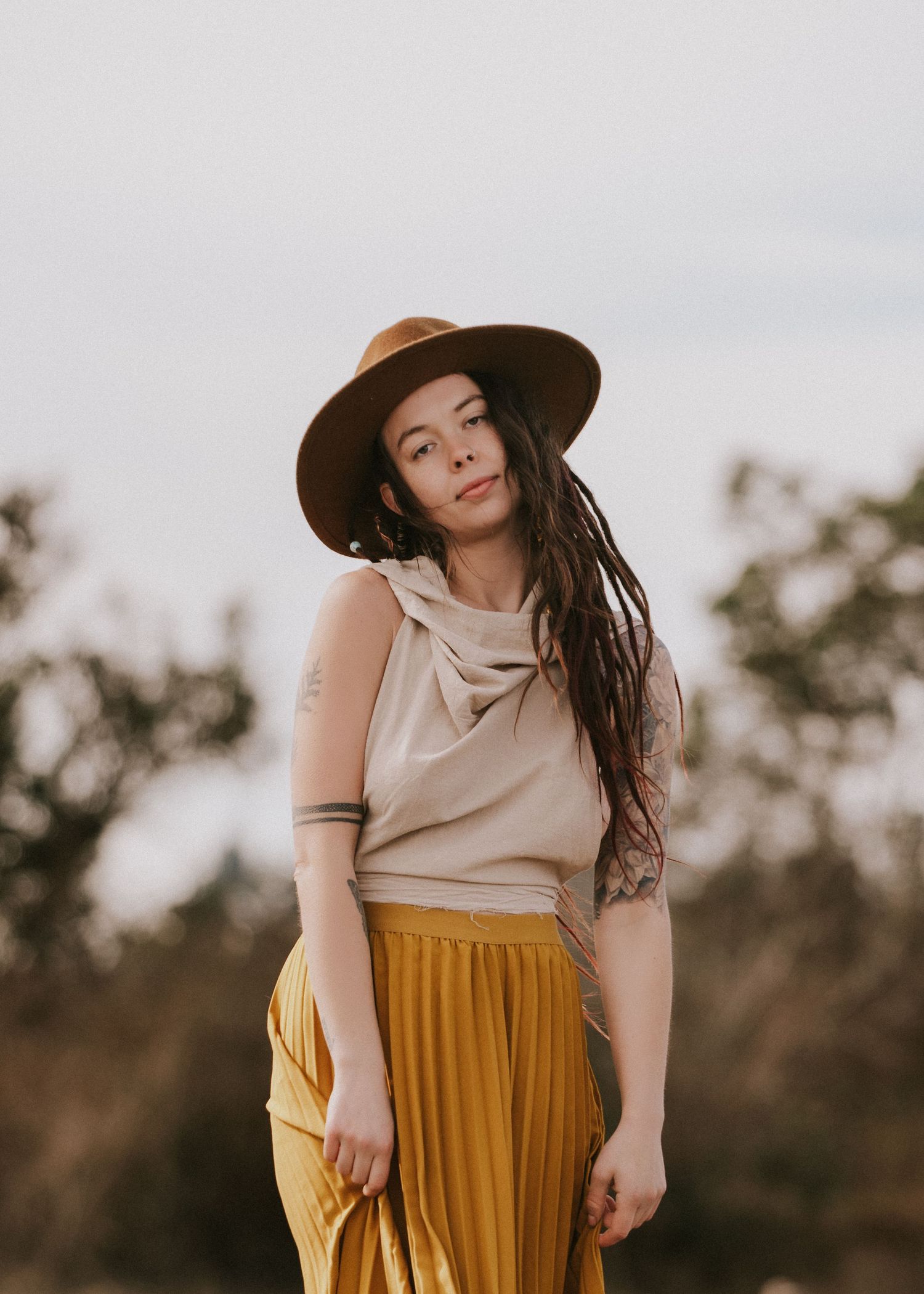 A woman in a mustard pleated skirt, beige top, and wide-brim hat poses outdoors with a bohemian style.