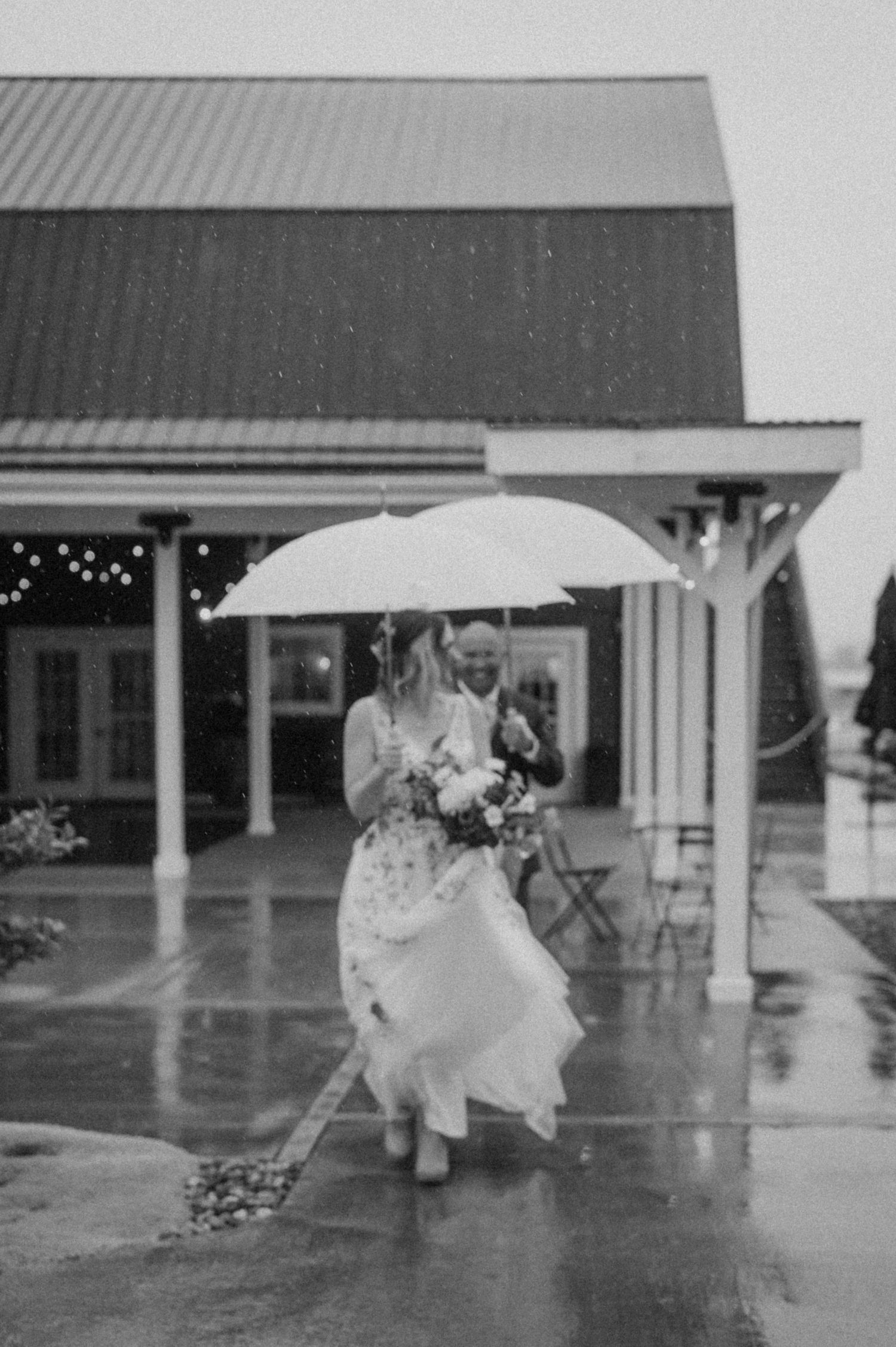 A rainy wedding day scene with white umbrella and flowing dress outside a rustic venue.