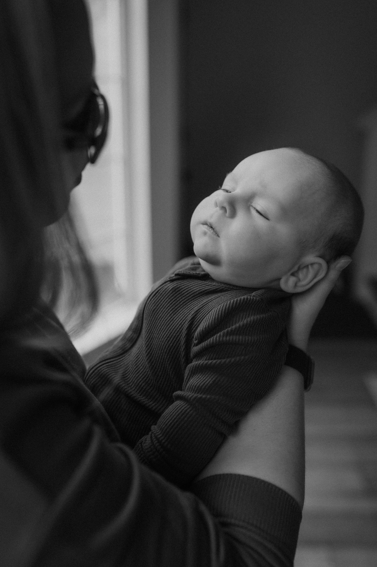 Black and white photo of a sleeping newborn baby cradled in an adult's arms near a window.