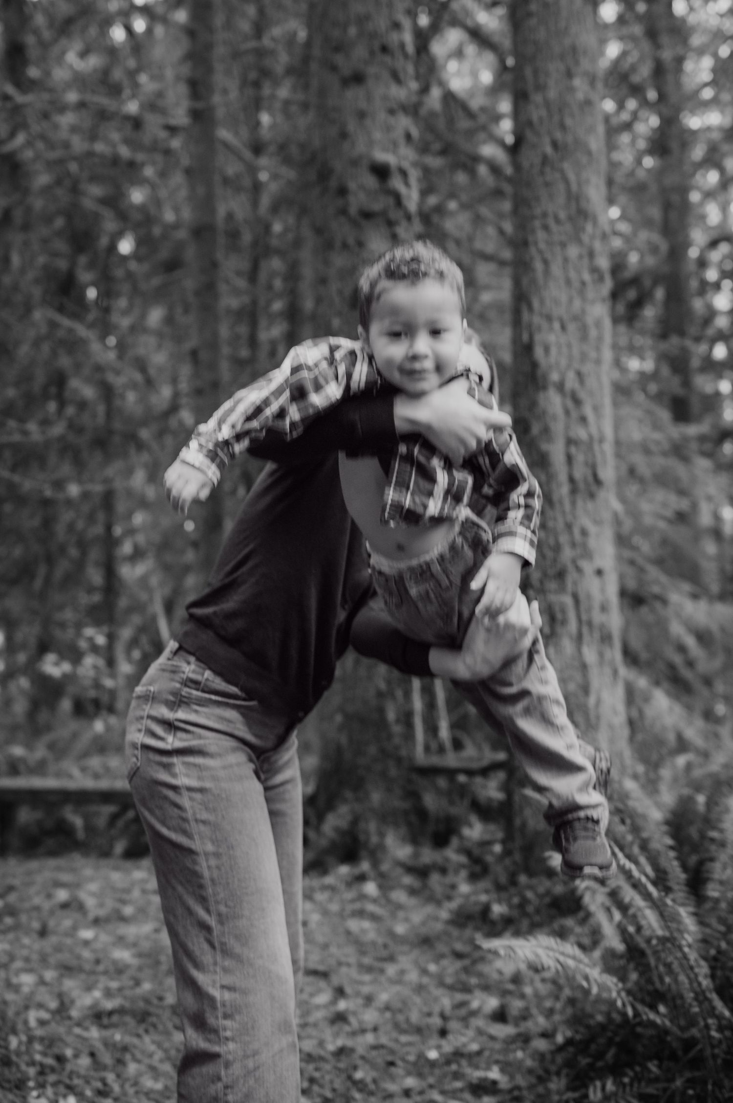 A child in a plaid shirt and jeans jumps playfully in a forest setting, captured in black and white.