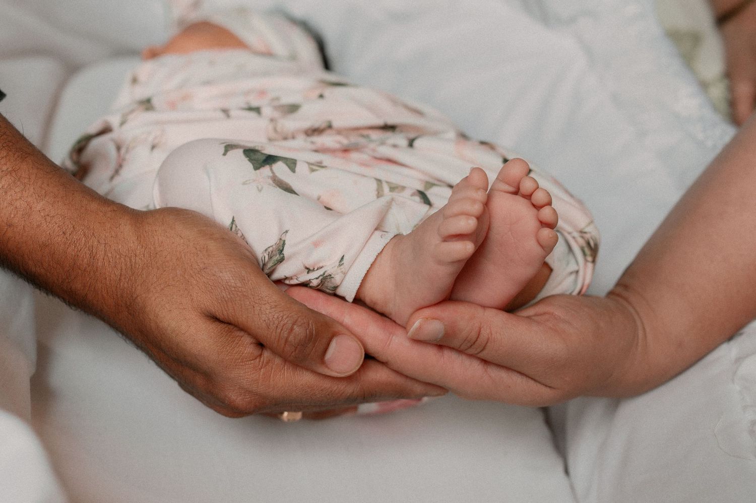 Hands of parents tenderly cradle a baby's feet wrapped in a floral pattern blanket on white bedding.