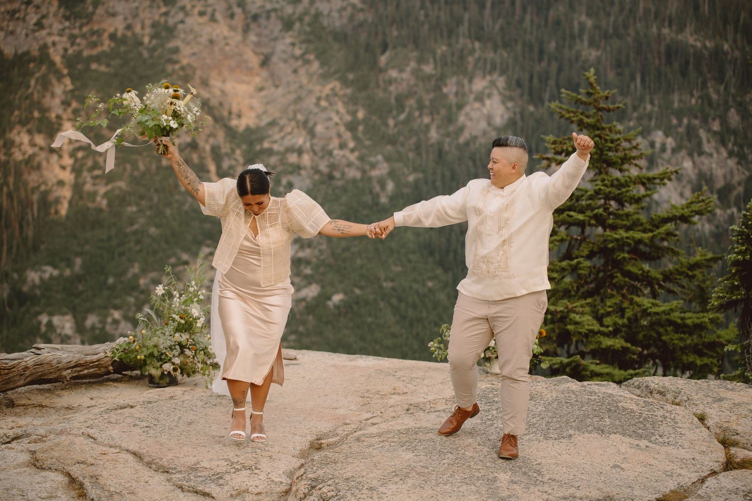 A couple in light colored outfits dances and celebrates together on a mountain cliff with dramatic scenery.