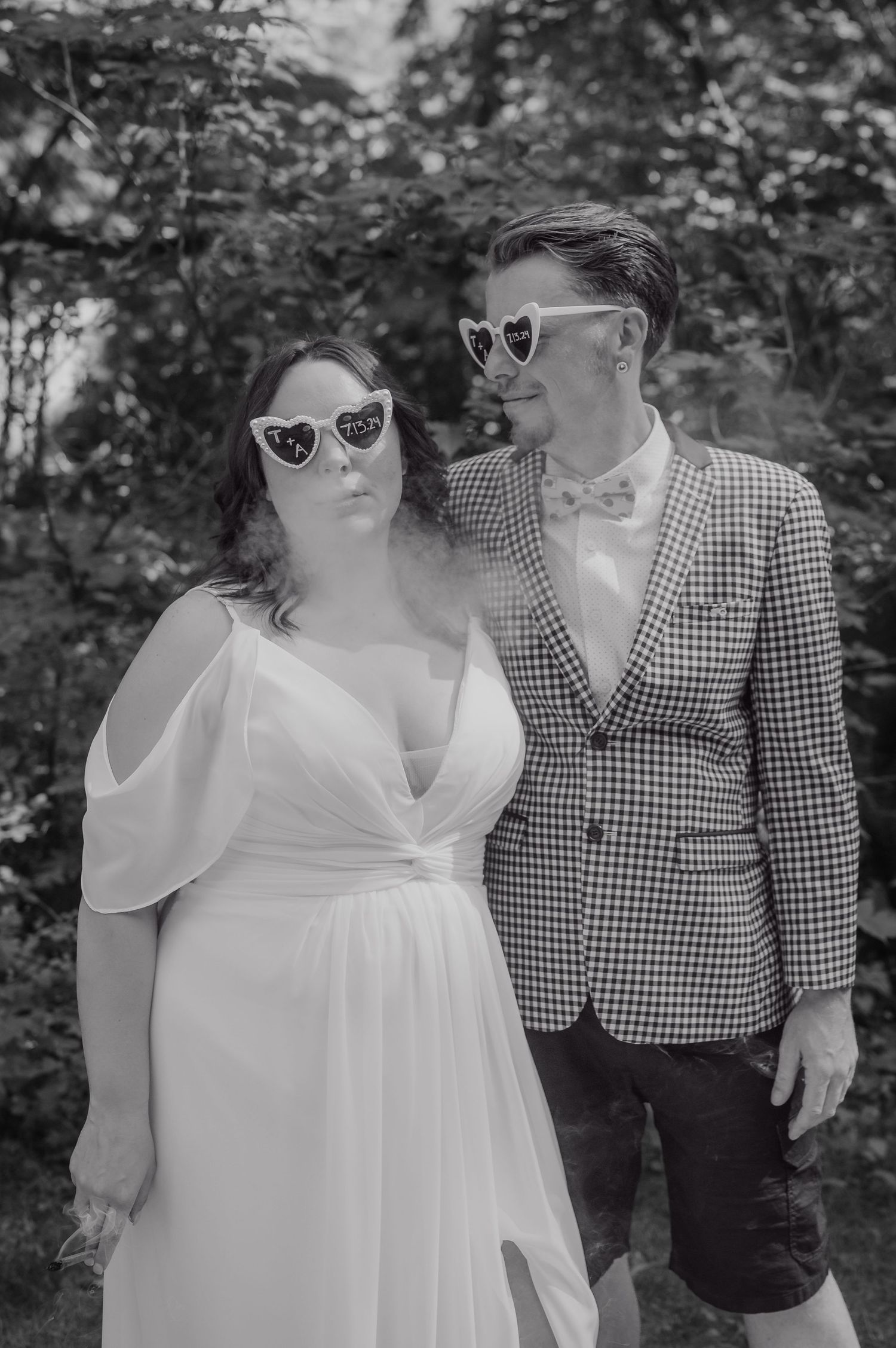 Two people in sunglasses pose together outdoors, one wearing a white dress and one in a checkered blazer and bowtie.
