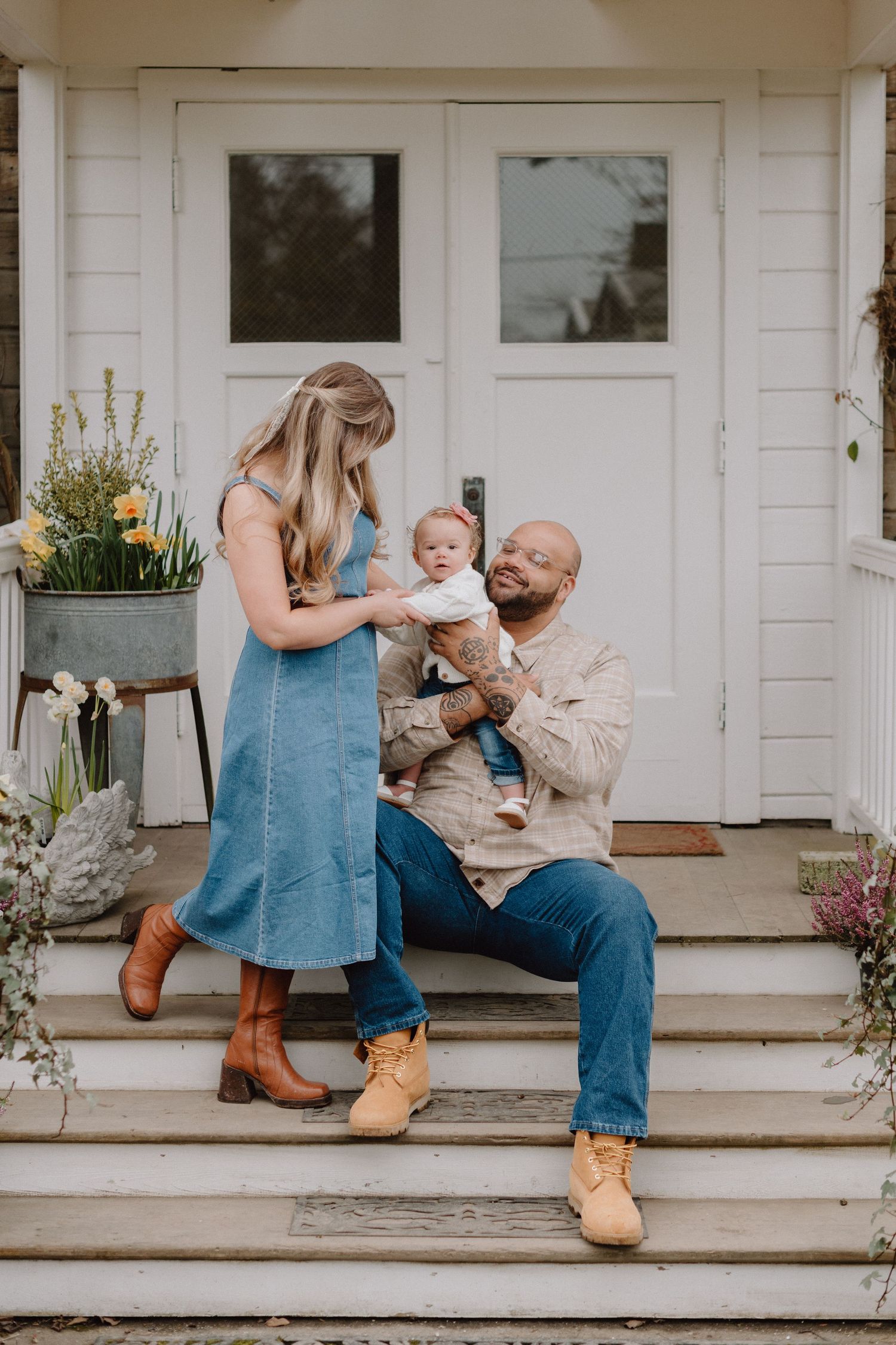 Young family with baby poses on white farmhouse porch steps surrounded by spring flowers.