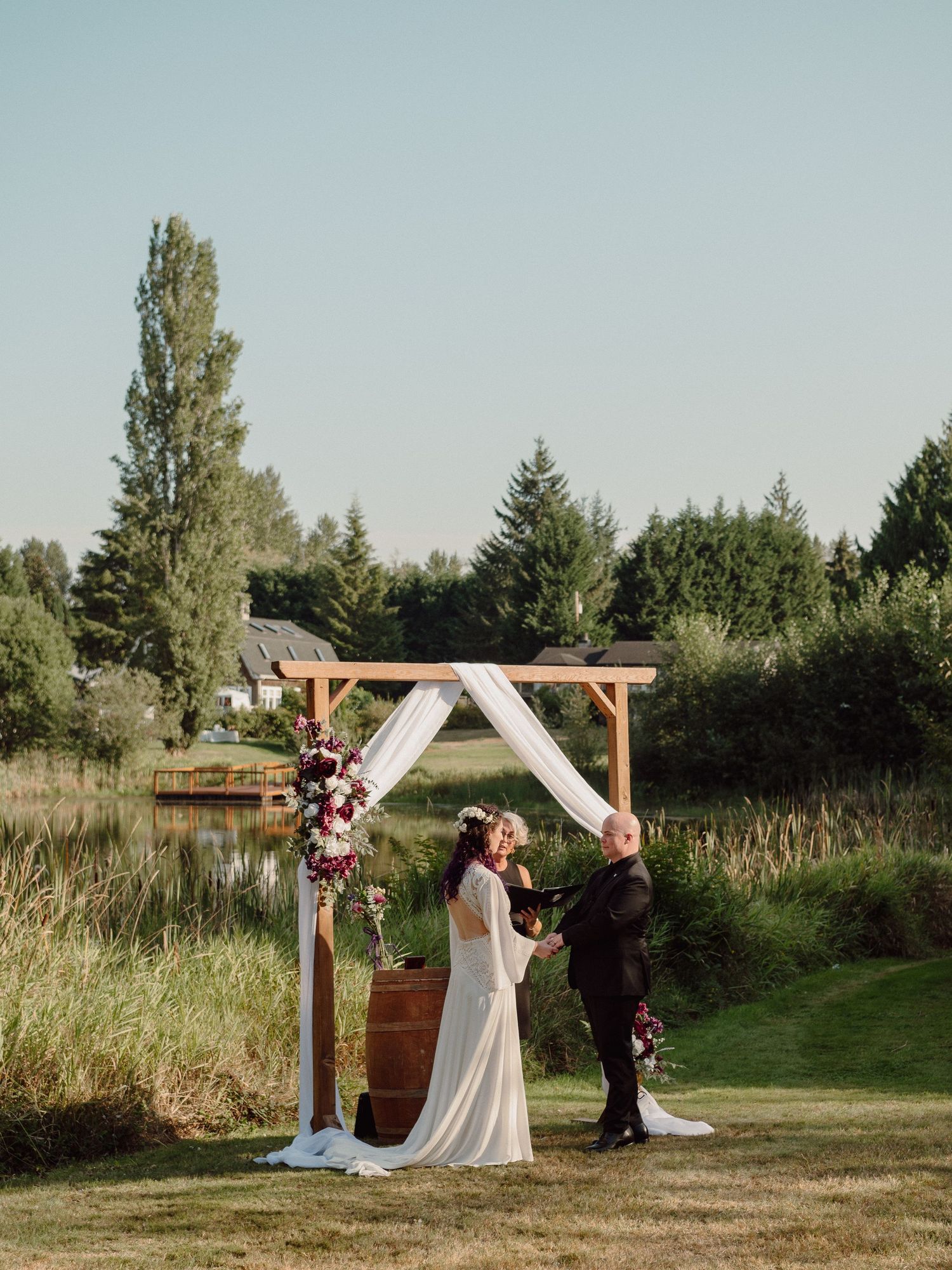 An outdoor wedding ceremony under a wooden arch decorated with flowers in a rustic garden setting.