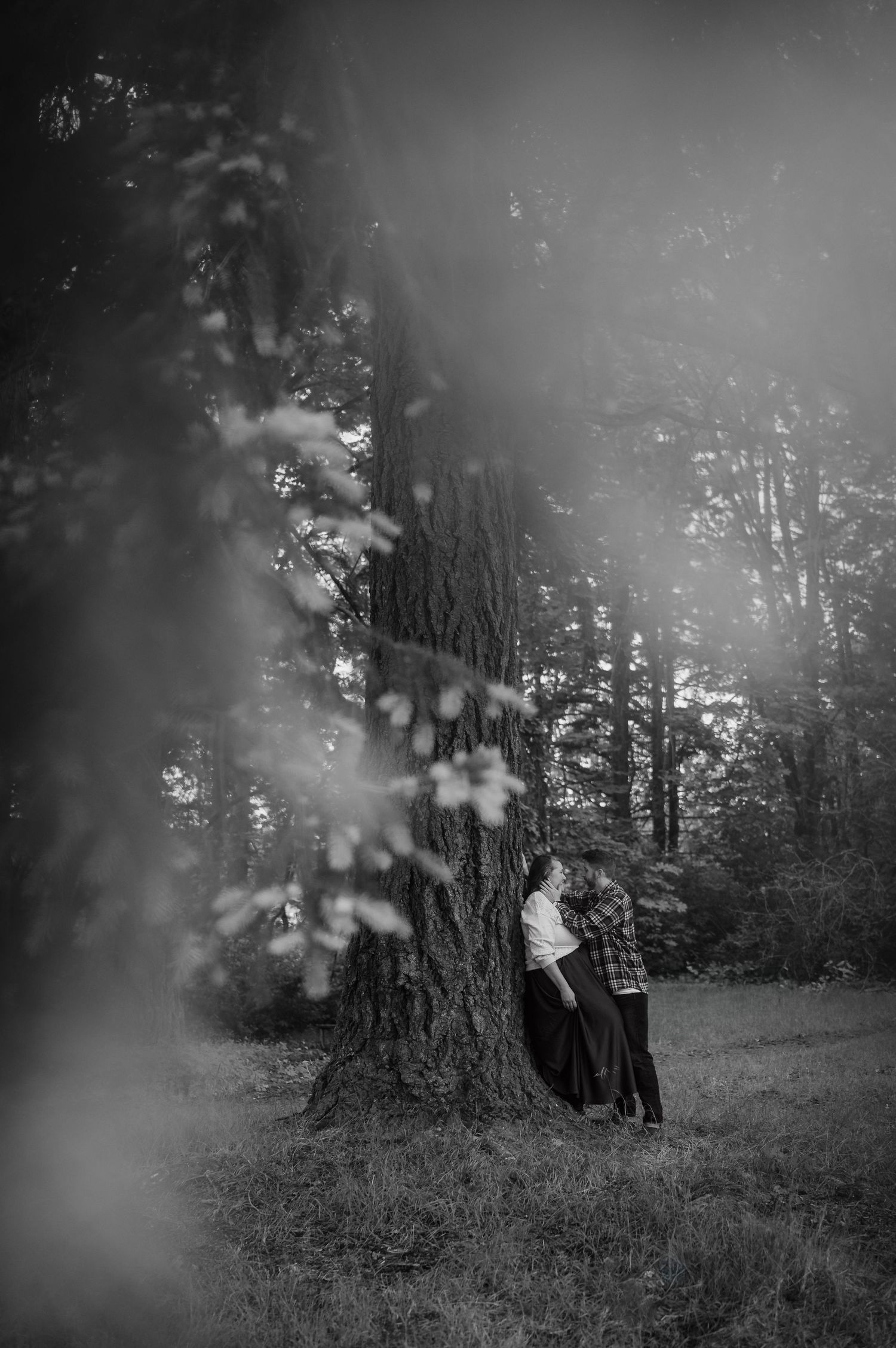 Black and white photo of a couple embracing beside a large tree in a misty forest setting.