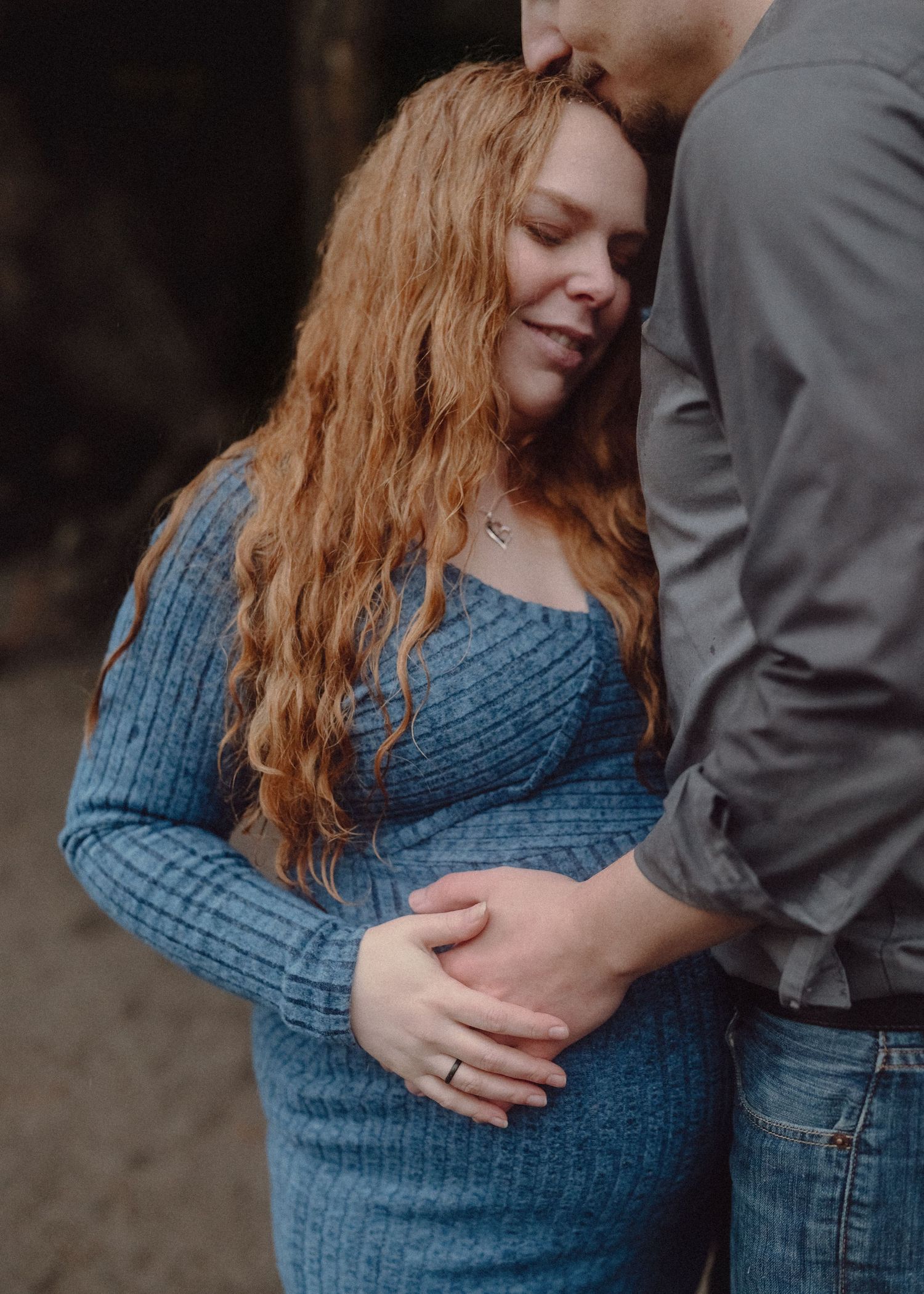 A pregnant woman in a blue ribbed dress cradles her baby bump while leaning against her partner outdoors.