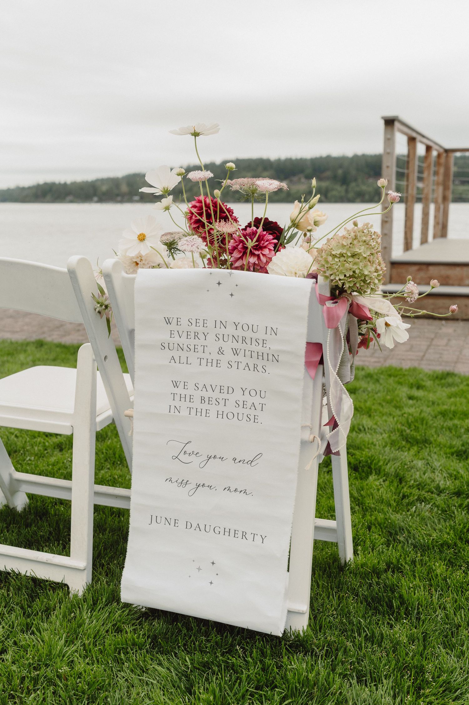 White wooden chairs with romantic wedding signs and floral decorations arranged on green grass by the waterfront.