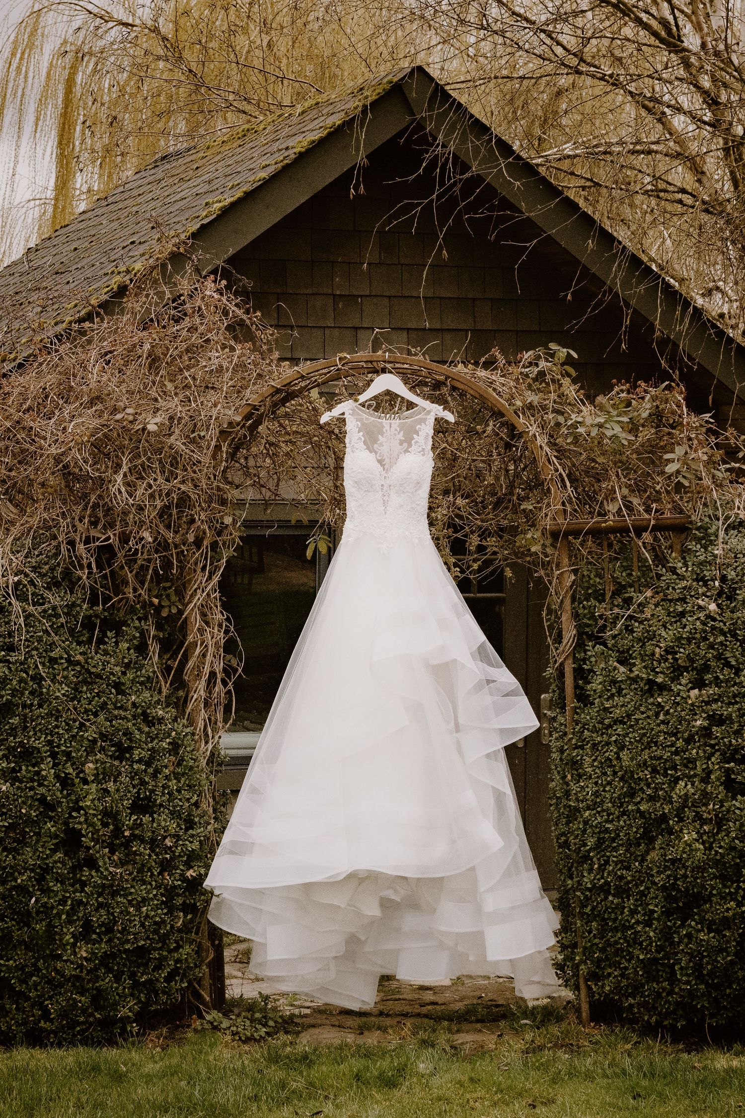 A white wedding dress hanging on an archway against a rustic wooden building with vines.