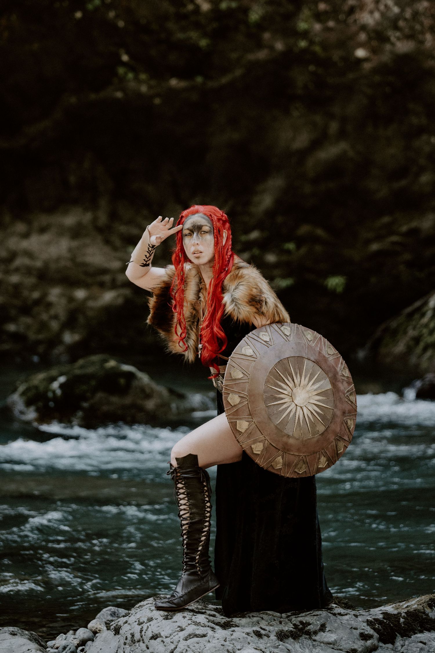 A warrior woman in Viking costume with red hair, fur cape, round shield, and tall boots stands on rocks by a river.