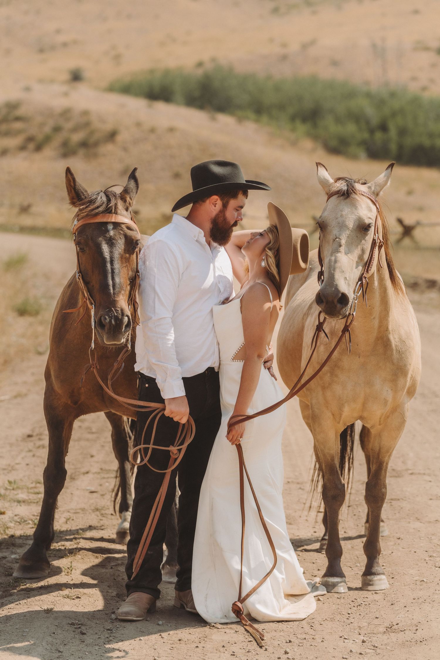 A couple with two horses posing together in a desert landscape wearing western attire.