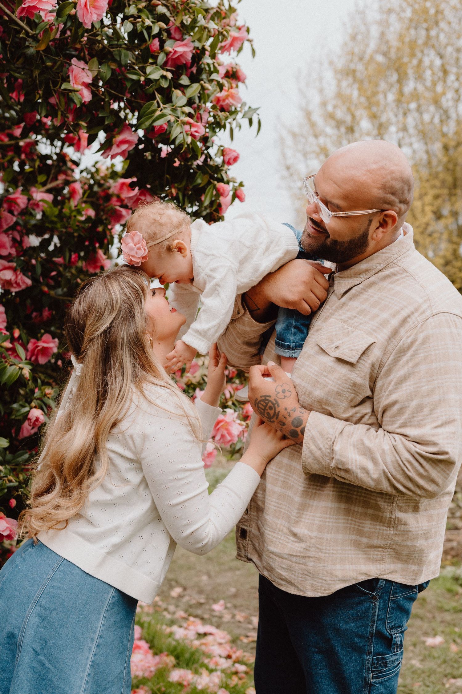 A family poses outdoors near pink blooming flowers, as a mother kisses her baby held by a tattooed bald father.