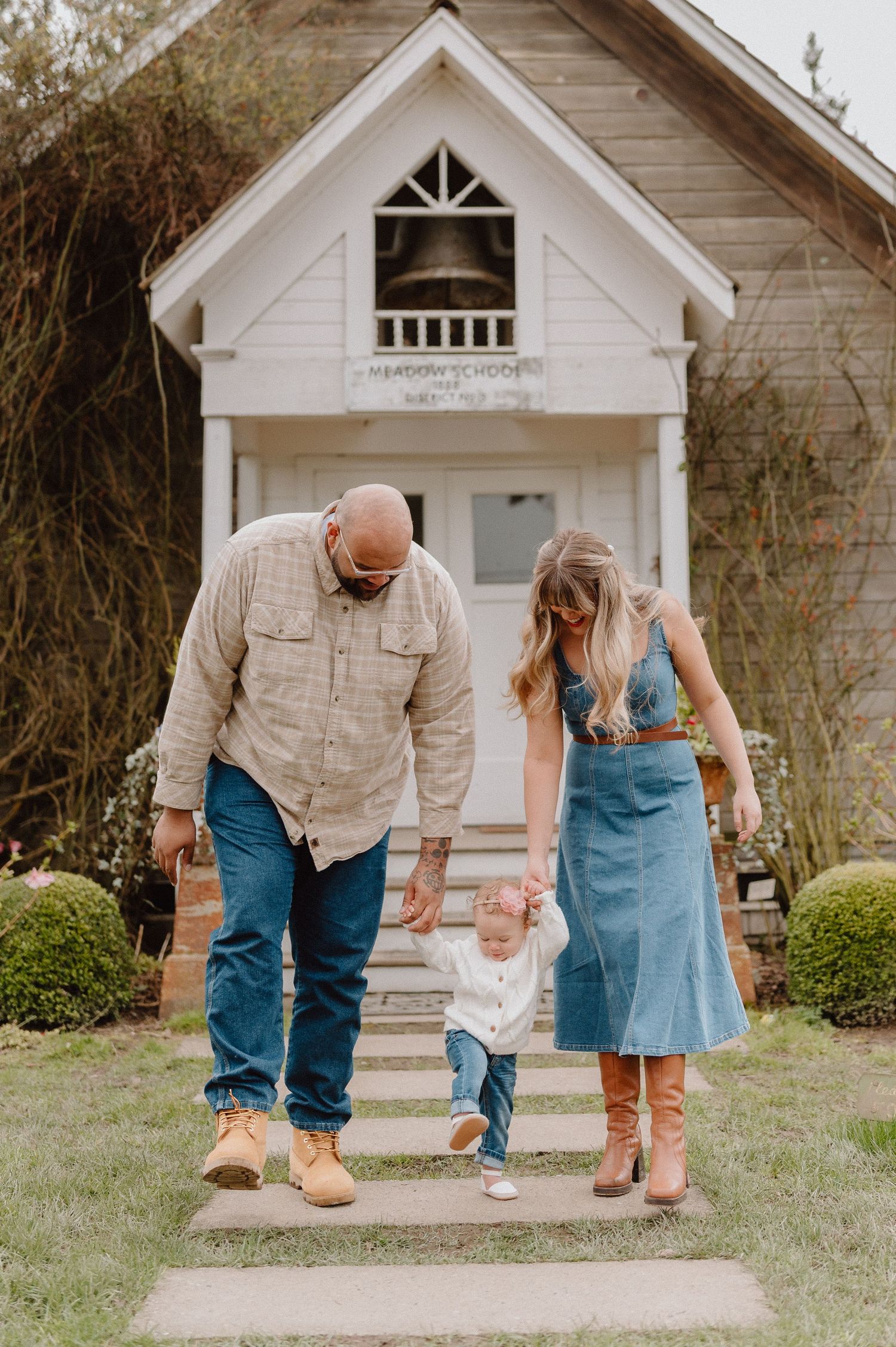 A family of three walks down steps outside a white chapel, parents holding their toddler's hands on a sunny day.