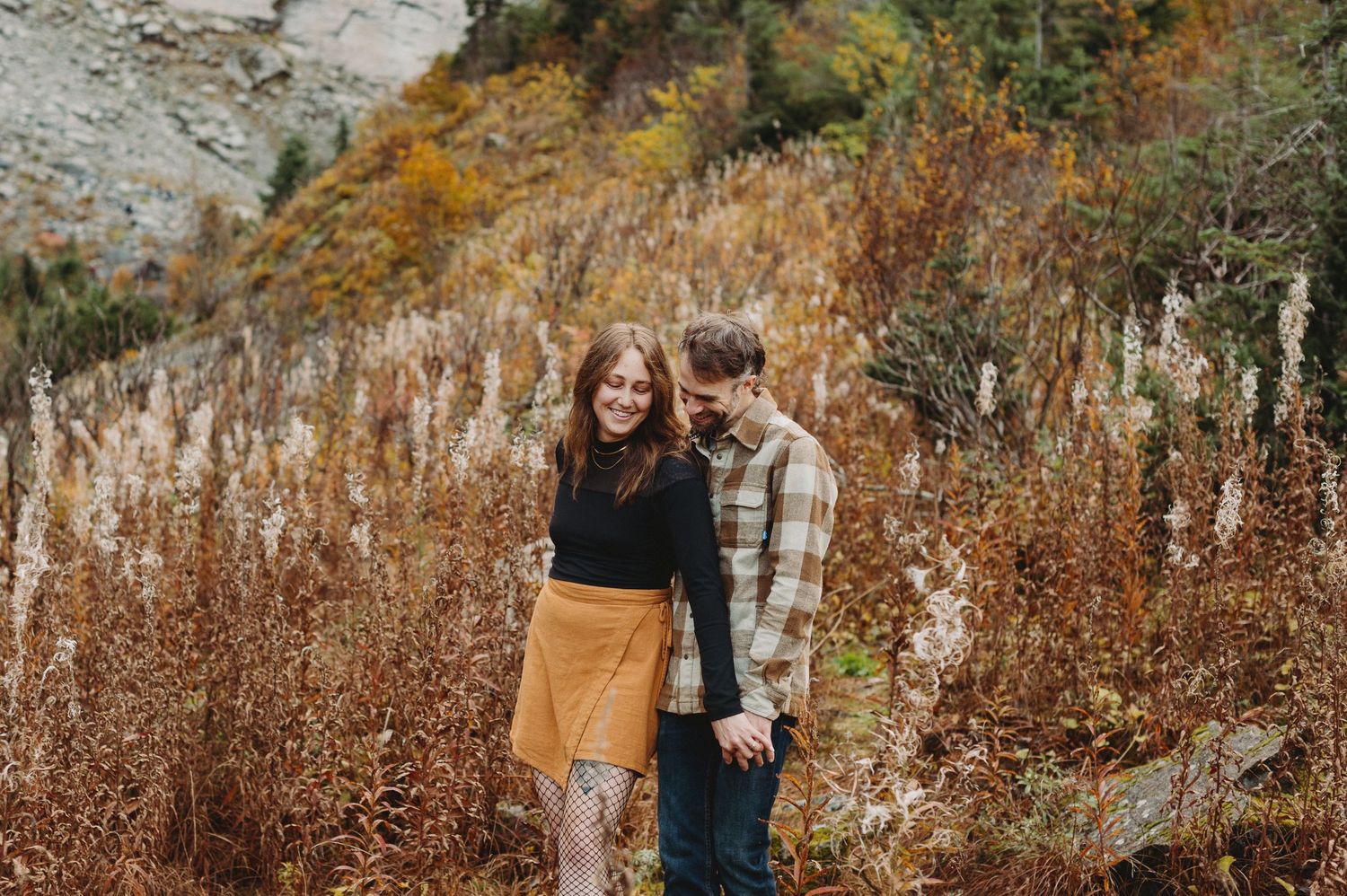 Couple walking through autumn foliage and tall grass in a scenic outdoor setting during golden hour.