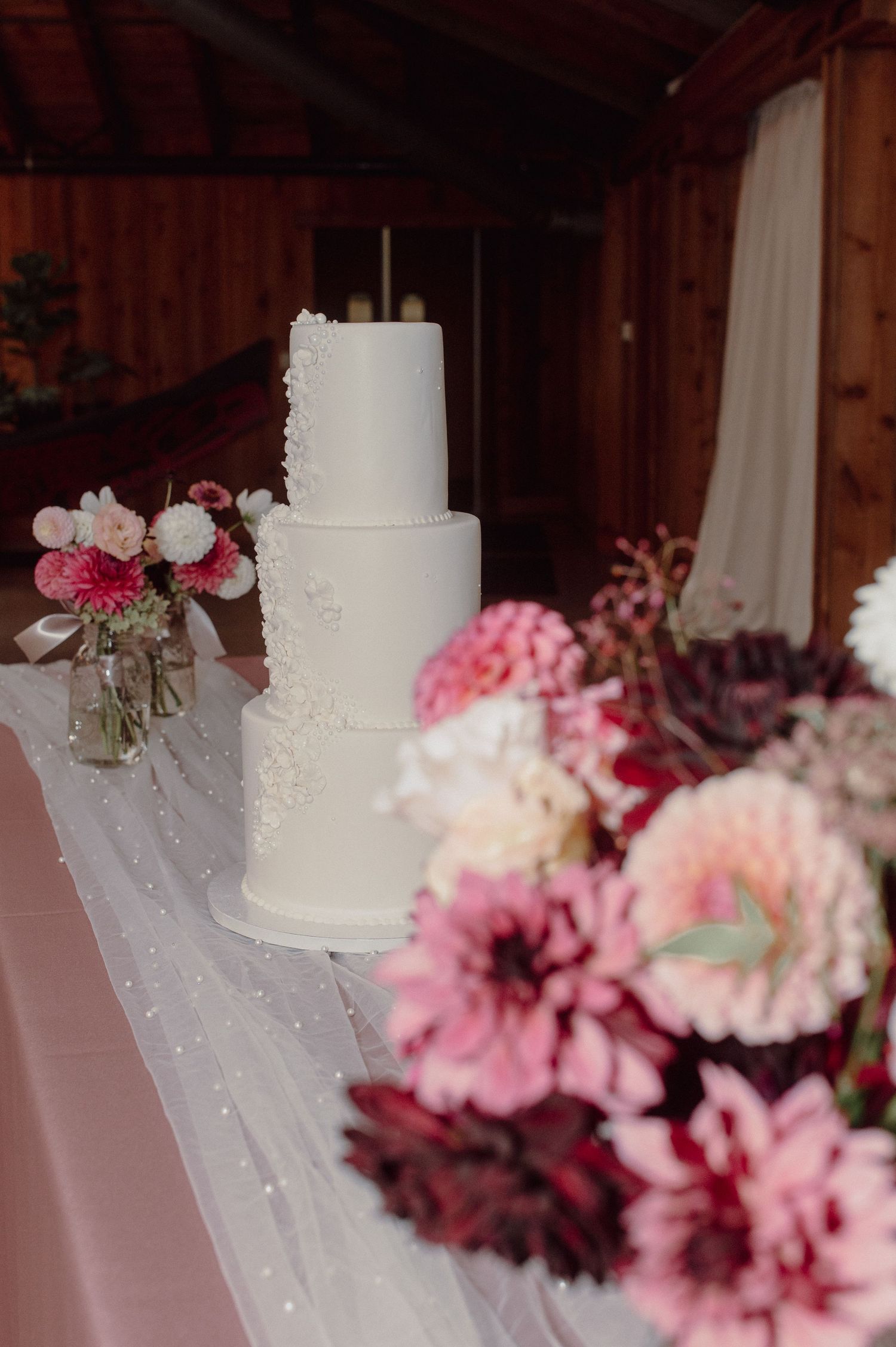 White tiered wedding cake decorated with delicate flowers and tulle against a backdrop of pink floral arrangements.